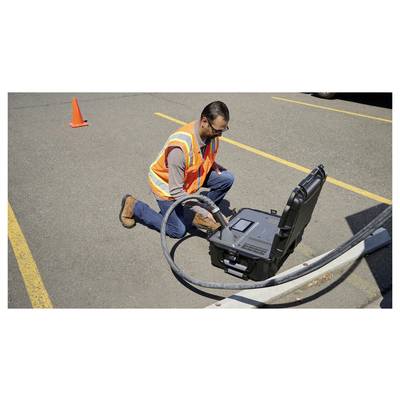 Une personne vêtue d'un gilet de sécurité s'agenouille sur la surface d'un parking à côté d'une caisse d'équipement ouverte, qui contient des dispositifs de surveillance électroniques.