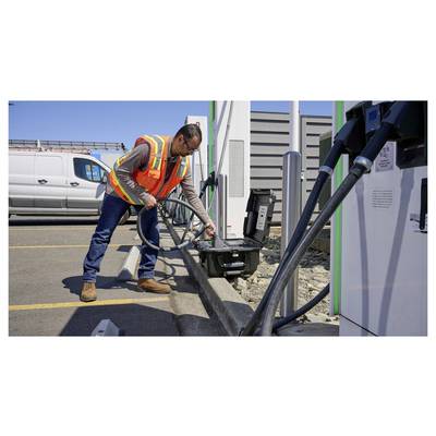 Une personne portant un gilet de sécurité branche un câble de recharge à un véhicule électrique à une station de recharge extérieure.