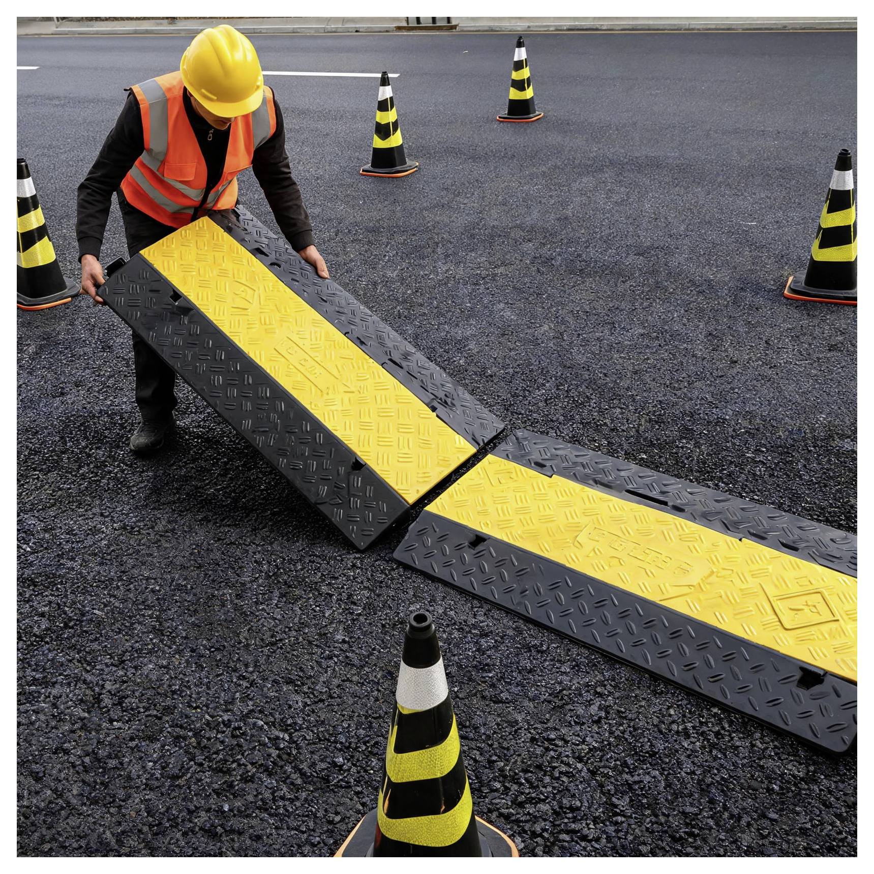 Travailleur vêtu d'un gilet de sécurité et d'un casque de protection installant une rampe de câble jaune et noir sur une route, entouré de cônes de signalisation.