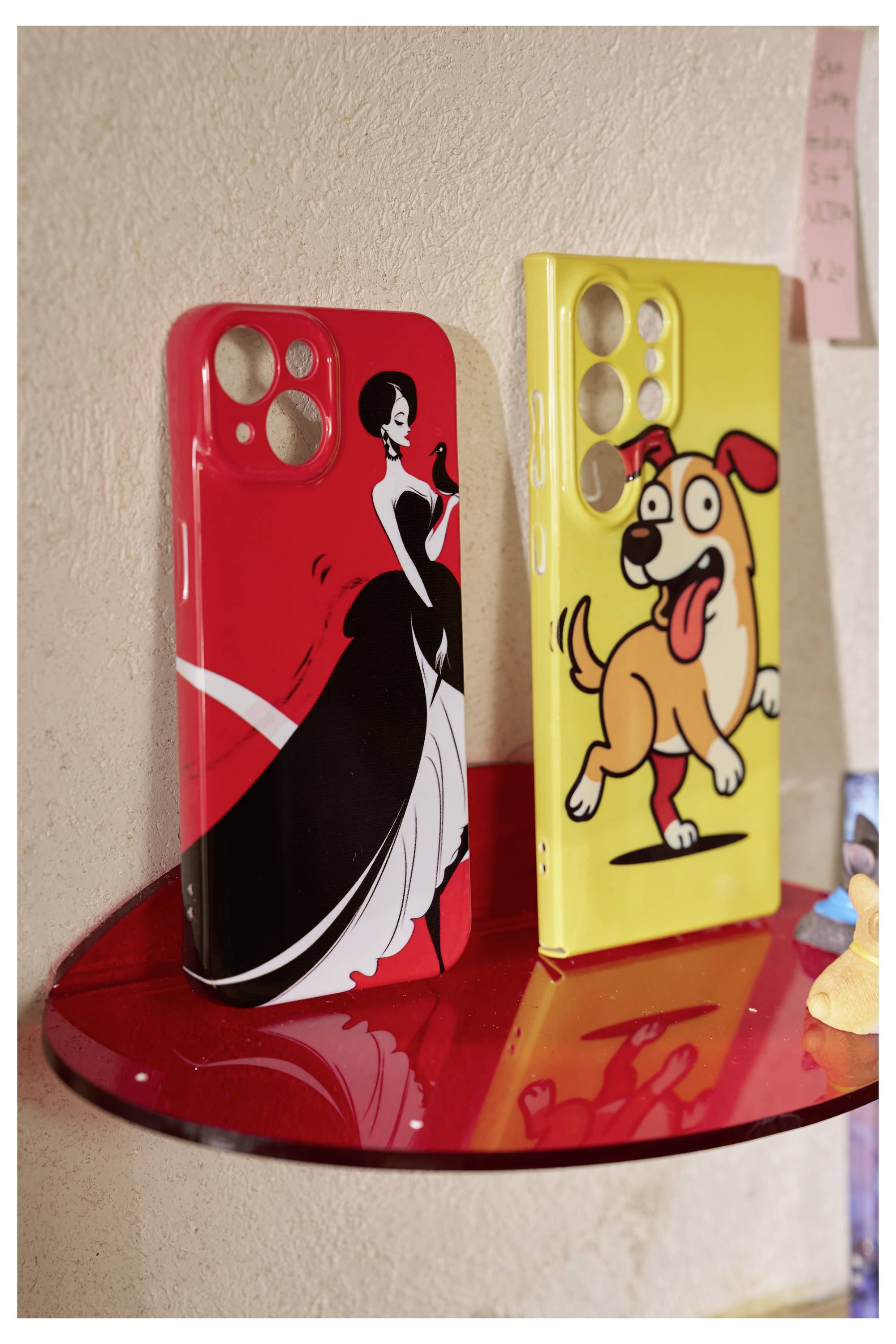 Deux étuis de téléphone sur une étagère rouge : l'un rouge avec une figure noire et blanche en robe, l'autre jaune mettant en vedette un chien de dessin animé.