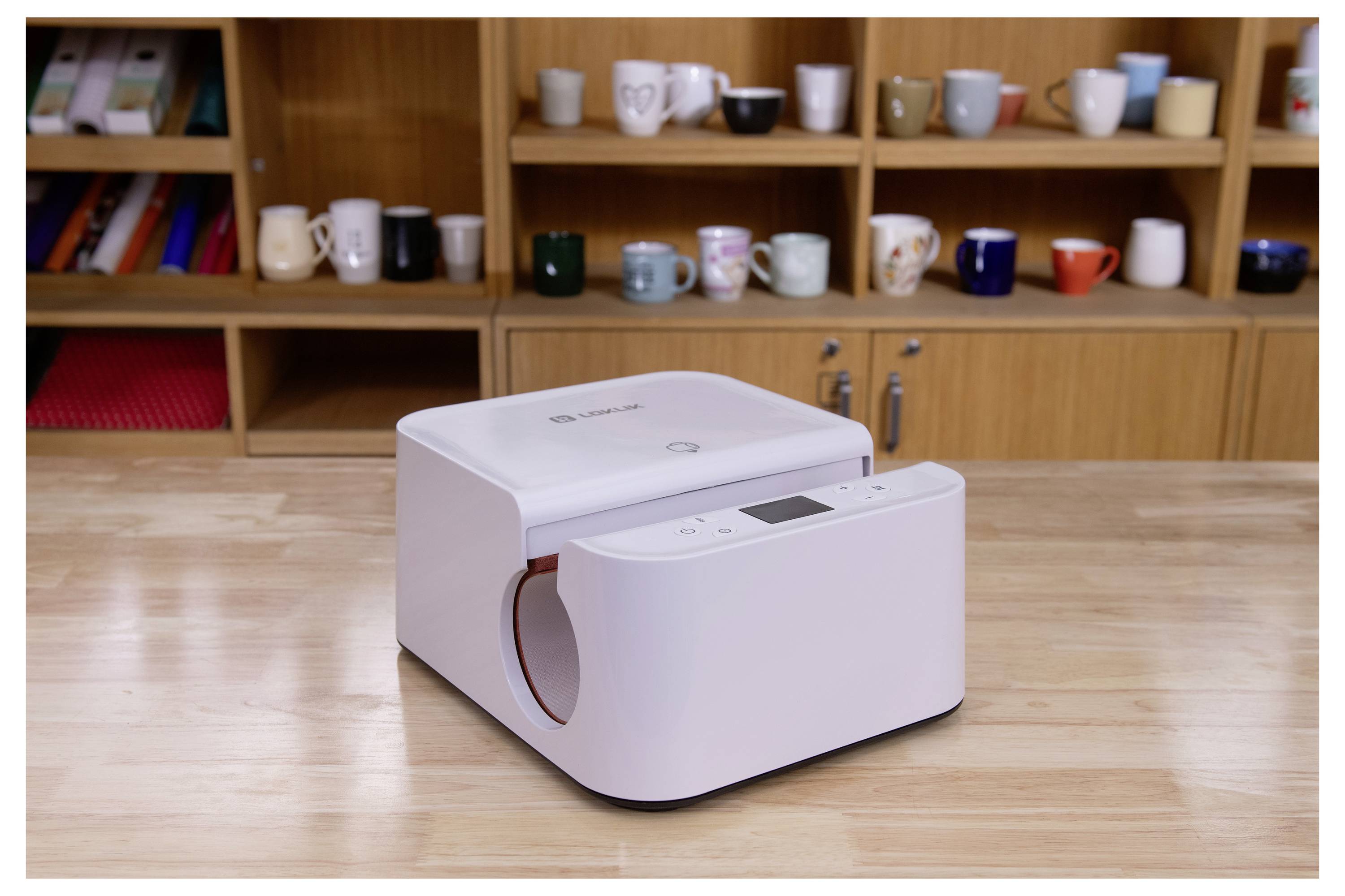 Un appareil de bureau au design épuré sur un bureau en bois, devant une étagère remplie de diverses tasses et gobelets.
