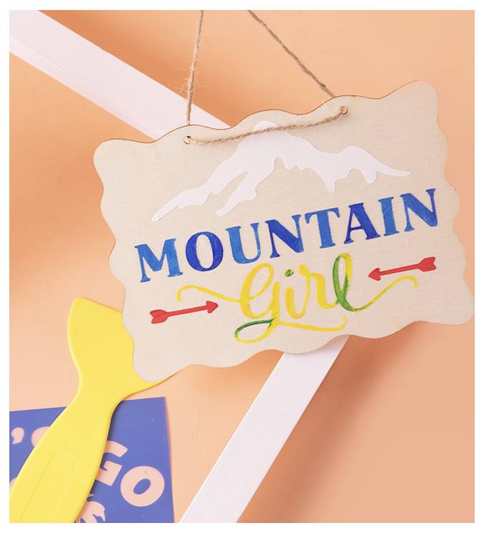 Une enseigne en bois avec le texte « MOUNTAIN Girl » en bleu et jaune, présentant des contours de montagnes et des flèches décoratives, suspendue sur un fond orange.