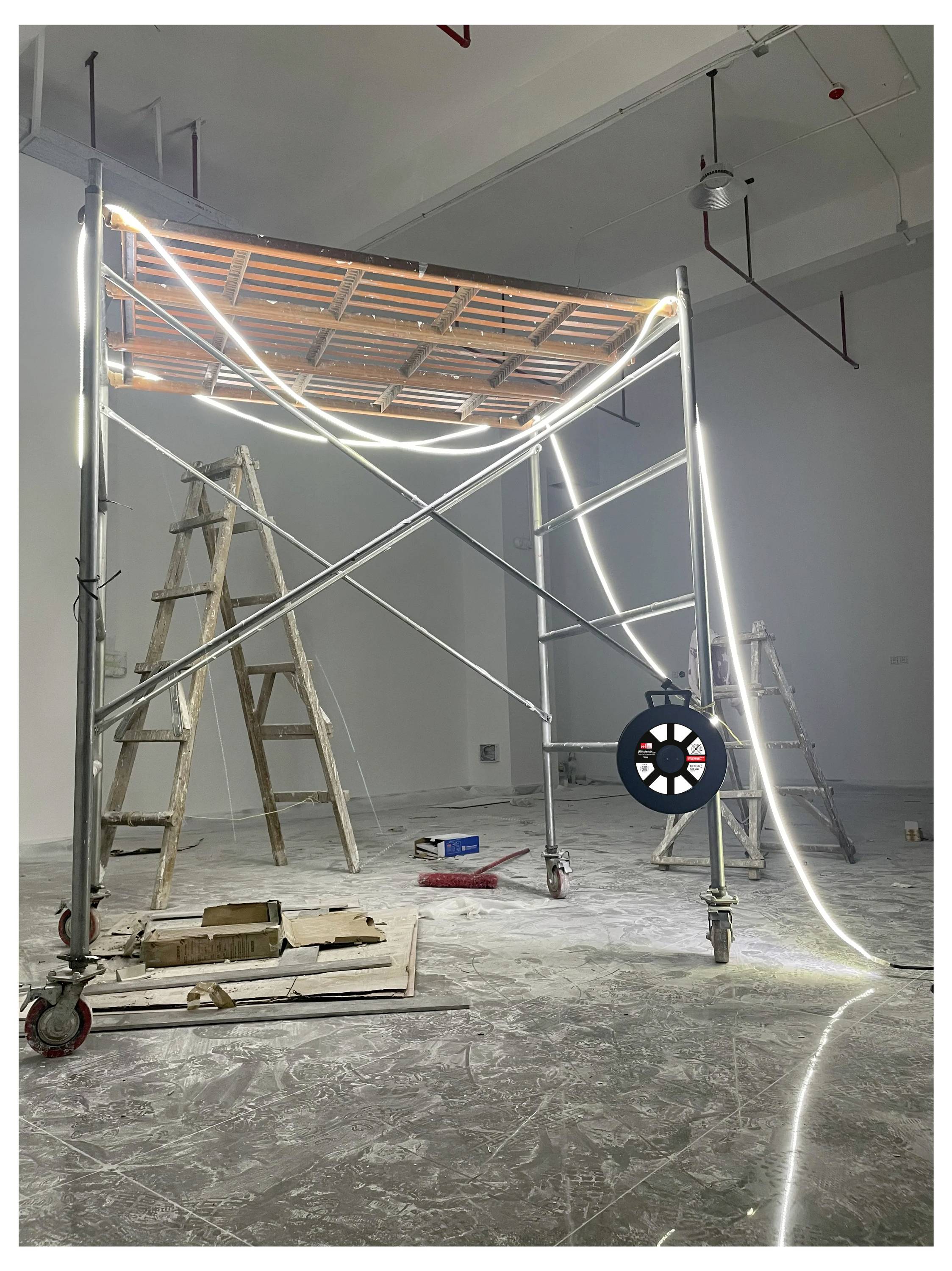 Installation artistique dans un espace galerie avec un échafaudage, éclairage par le haut et autour de la base, entourée d'escabeaux et d'outils dispersés sur le sol.
