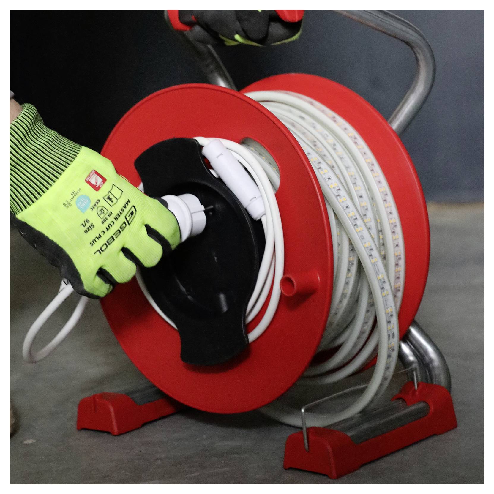 Une personne portant des gants verts déroule un câble électrique blanc à partir d'une bobine de câble rouge posée sur un sol gris.