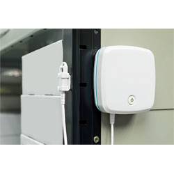 Un appareil électronique blanc monté sur un mur, connecté via un câble, possiblement un capteur ou une alarme, avec des étagères en arrière-plan.