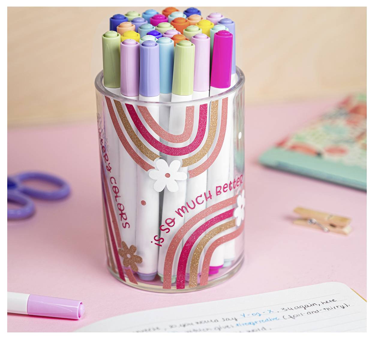 Une tasse transparente remplie de stylos colorés assortis présentant un design arc-en-ciel et fleurs, et le texte « tellement mieux » dans une police ludique.