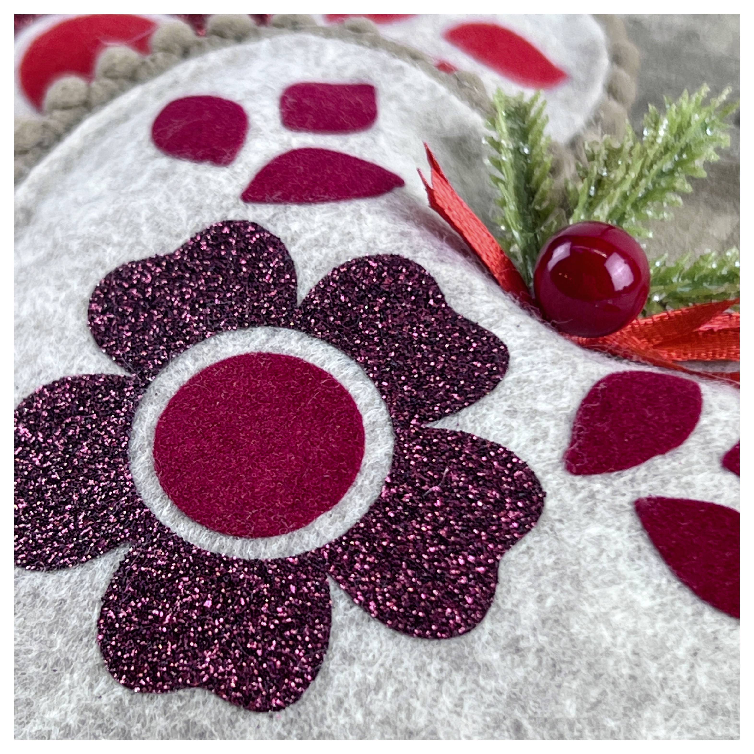 Un gros plan d'une décoration de vacances festive présentant un design de fleur pailletée sur feutre, avec des accents rouges et du faux feuillage.