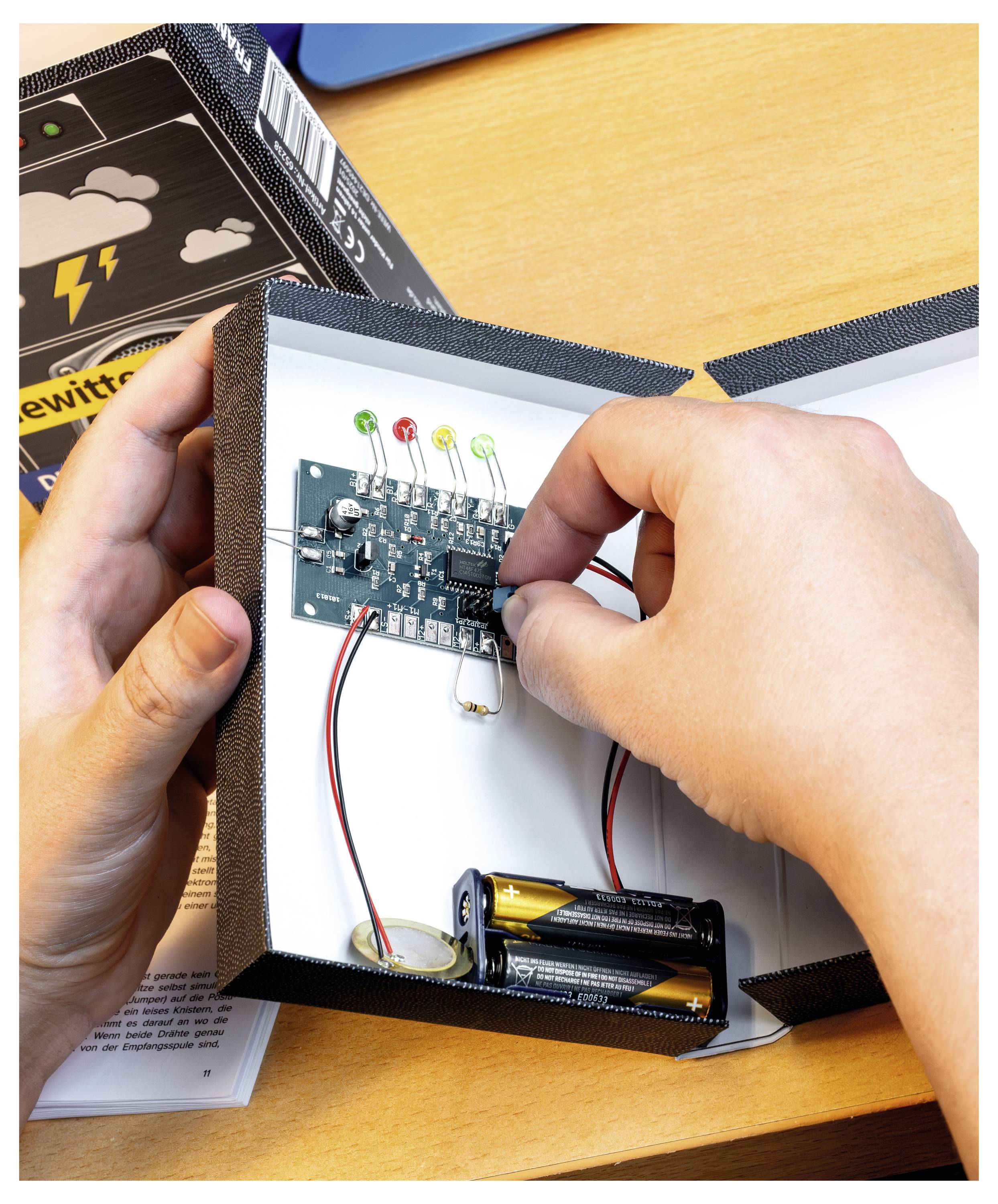Les mains ajustent une carte de circuit à l'intérieur d'une boîte ; une batterie et des LED sont visibles. Un livre intitulé « lewit » et un diagramme d'éclair sont partiellement visibles.