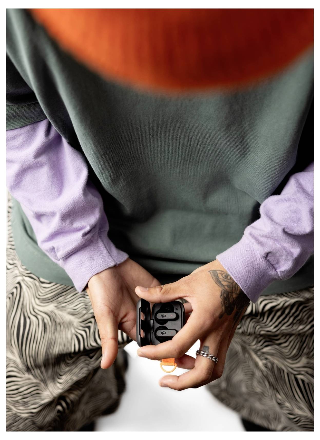 Une personne vêtue d'un t-shirt vert, de manches longues violettes, d'un bonnet tricoté orange et d'un pantalon à motifs tient une mallette noire avec des écouteurs, regardant vers le bas.