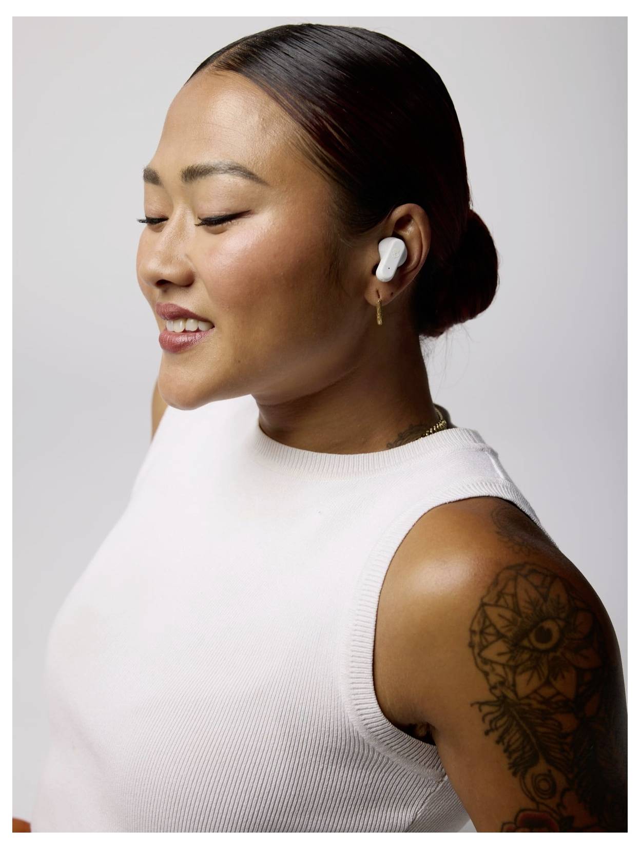 Une personne avec des écouteurs sourit, portant un haut blanc sans manches. Son bras a un tatouage, et les cheveux sont coiffés en un chignon soigné.