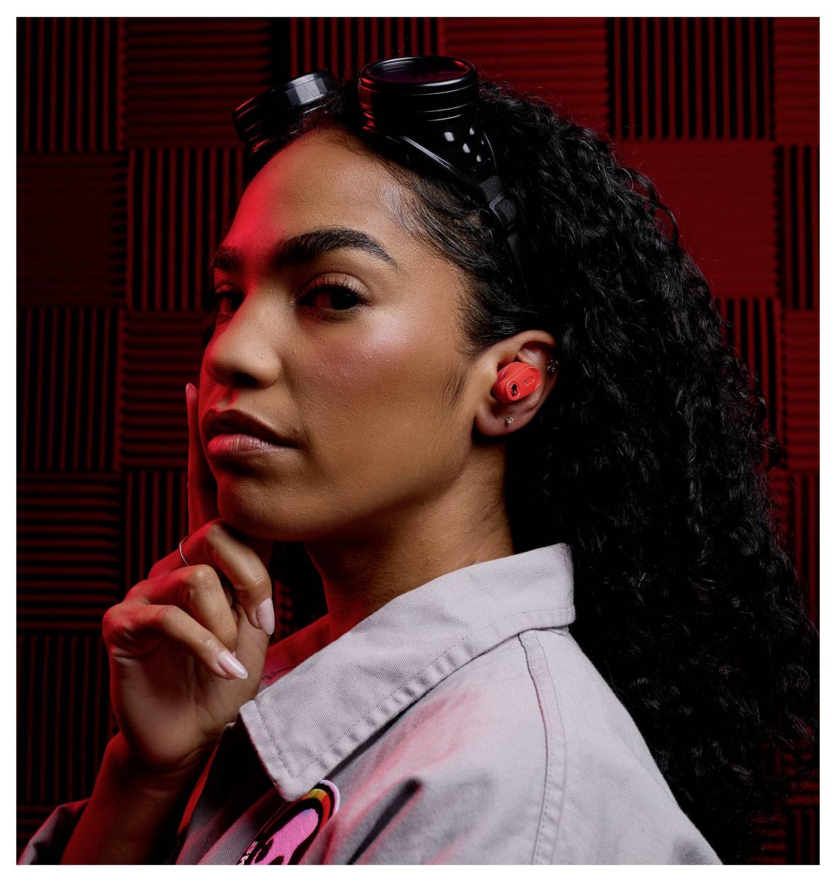 Une femme vêtue d'une chemise grise avec un patch coloré sur l'épaule, portant des lunettes noires sur sa tête, pose avec une expression pensive.