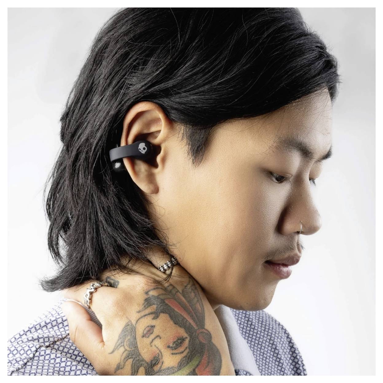 Une personne avec des écouteurs, portant une chemise motif. Elle a les cheveux noirs longs, un piercing au nez et un tatouage sur la main.