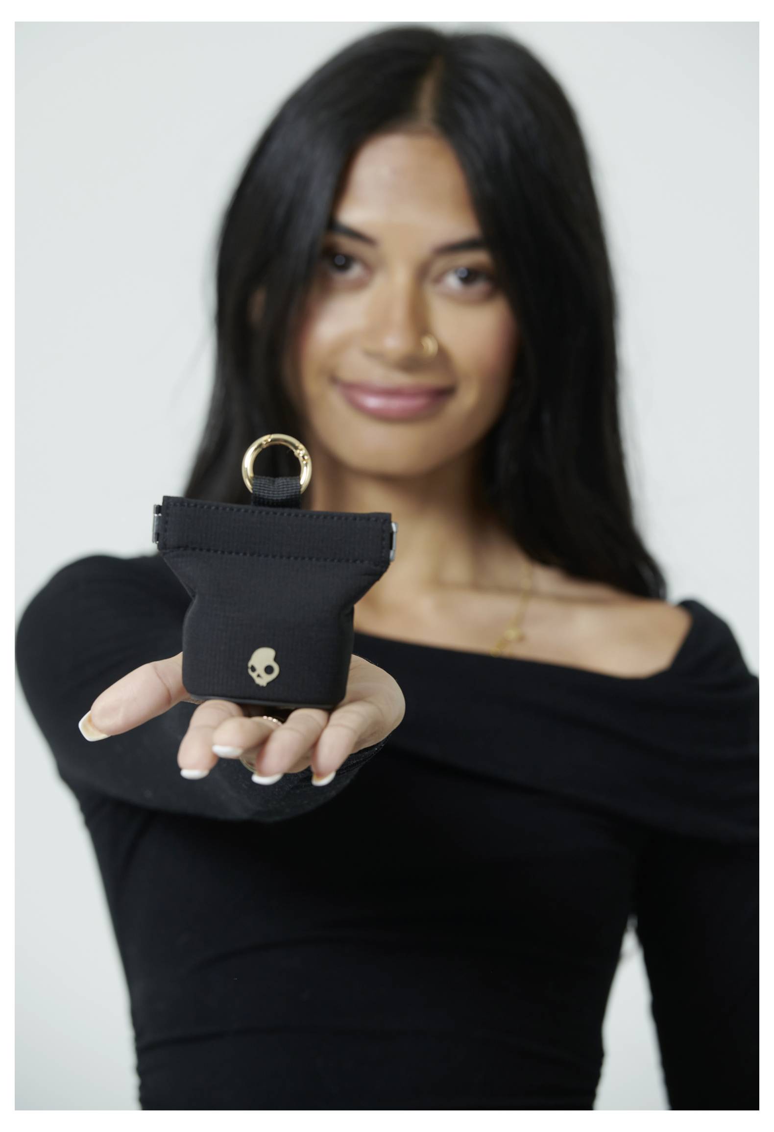 Une femme vêtue d'un haut noir tient un petit sac noir avec un anneau doré et un emblème de crâne, tendu vers l'appareil photo, en souriant.