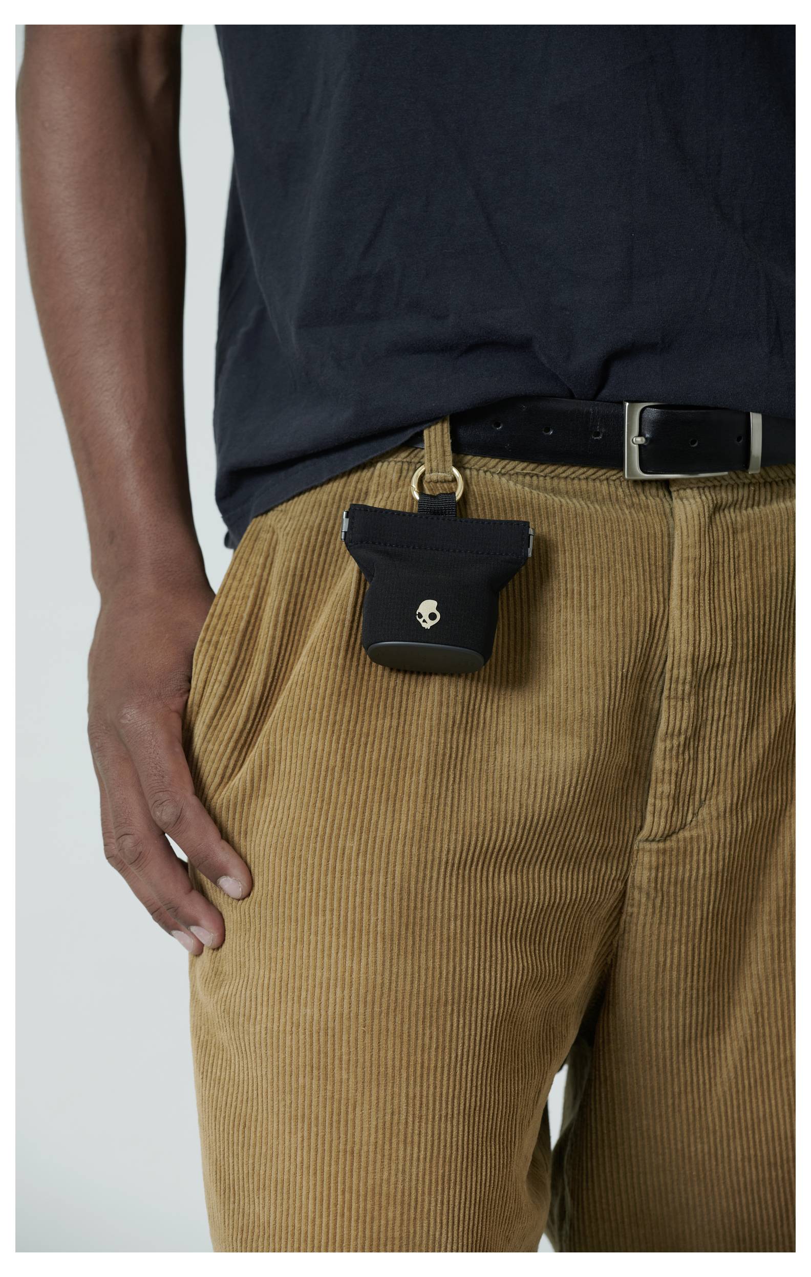 Une personne portant un pantalon en velours côtelé marron et une chemise noire, avec une petite pochette noire ornée d'un motif de crâne attachée à la ceinture.