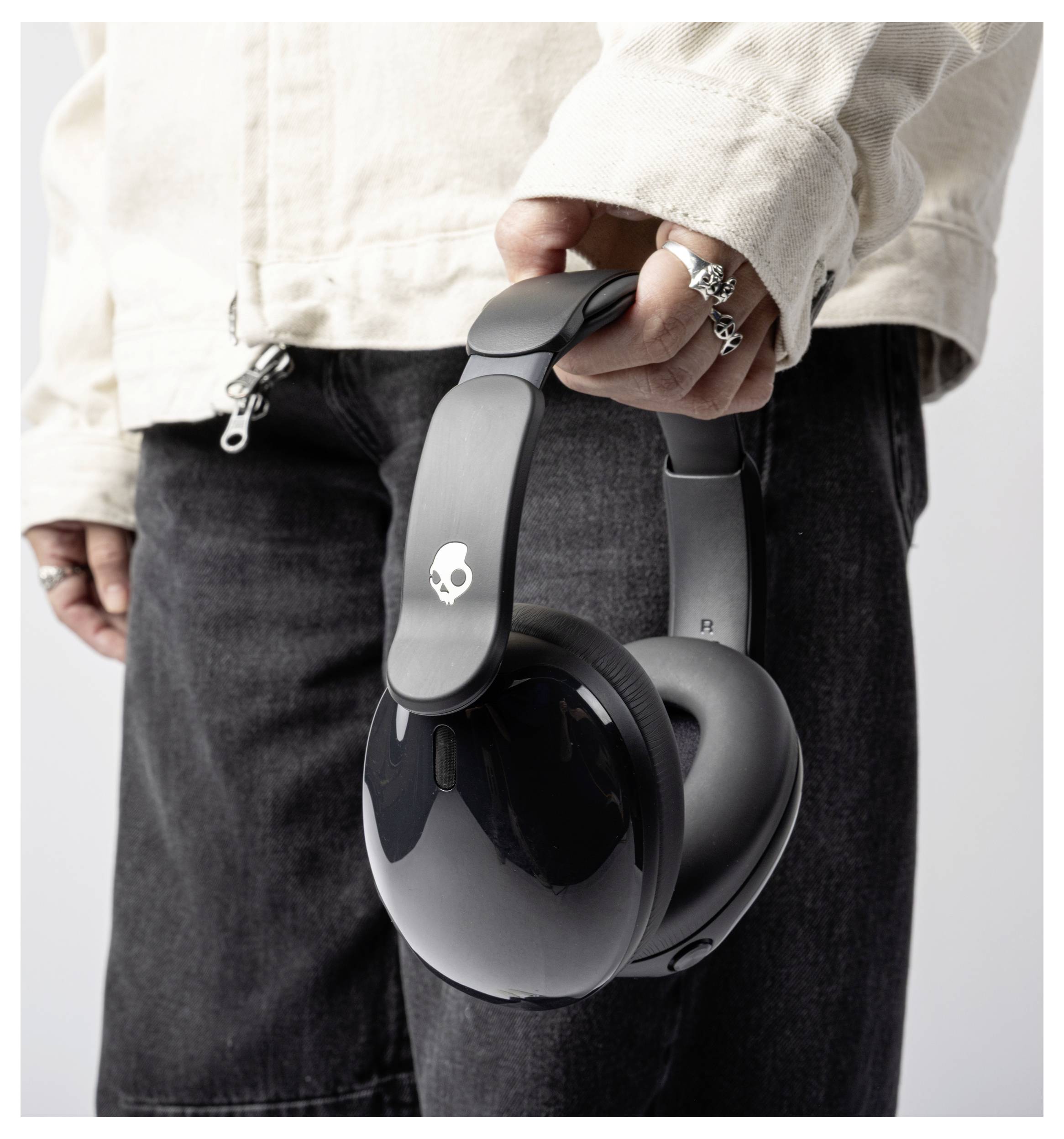 Une personne portant une veste beige tient une paire de casques supra-auriculaires noirs avec un logo de crâne, vêtue d'un jean foncé, mettant en avant un style décontracté.