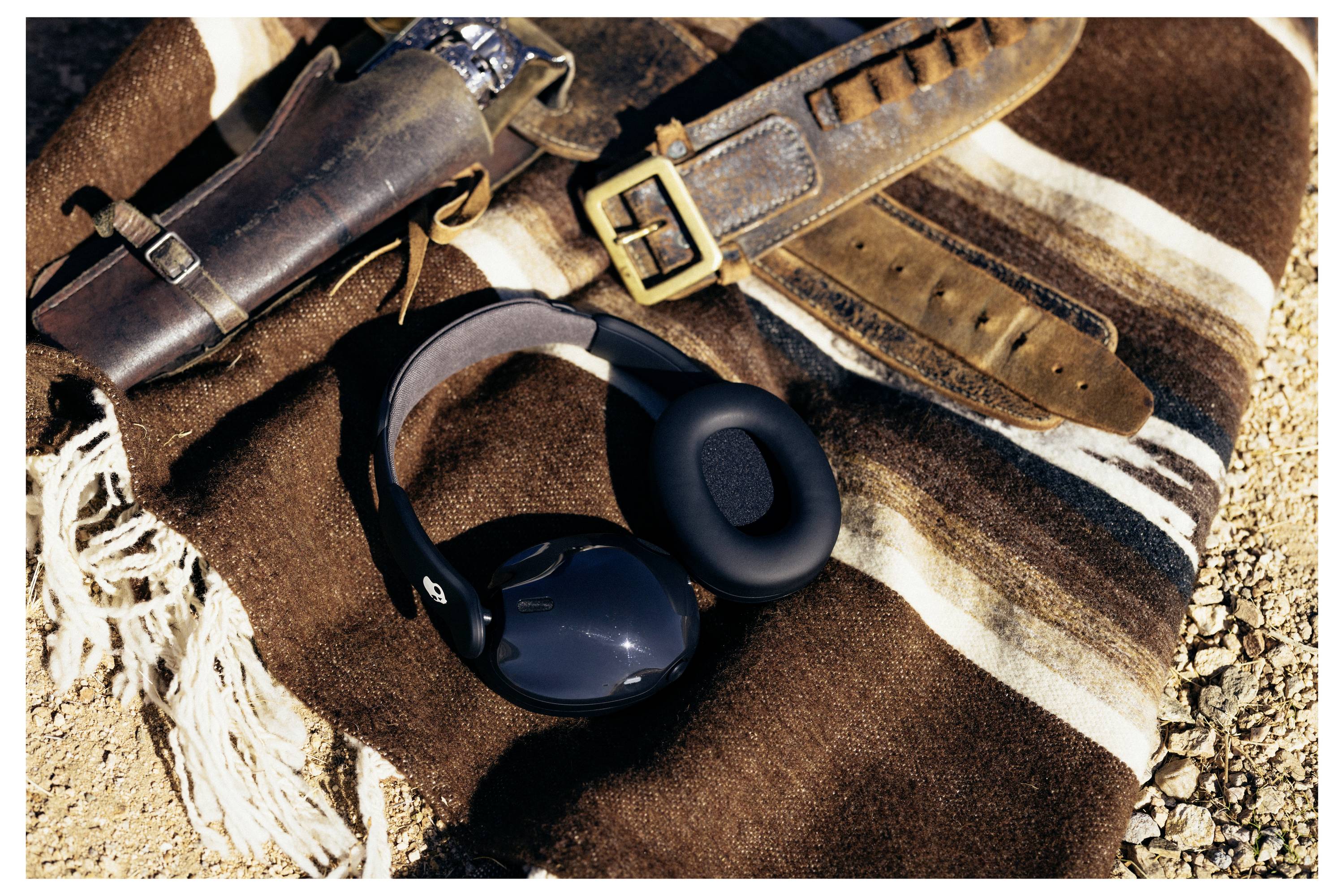 « Casque audio reposant sur une couverture rayée à côté d'un étui à pistolet en cuir marron et d'une ceinture dans un décor désertique. »