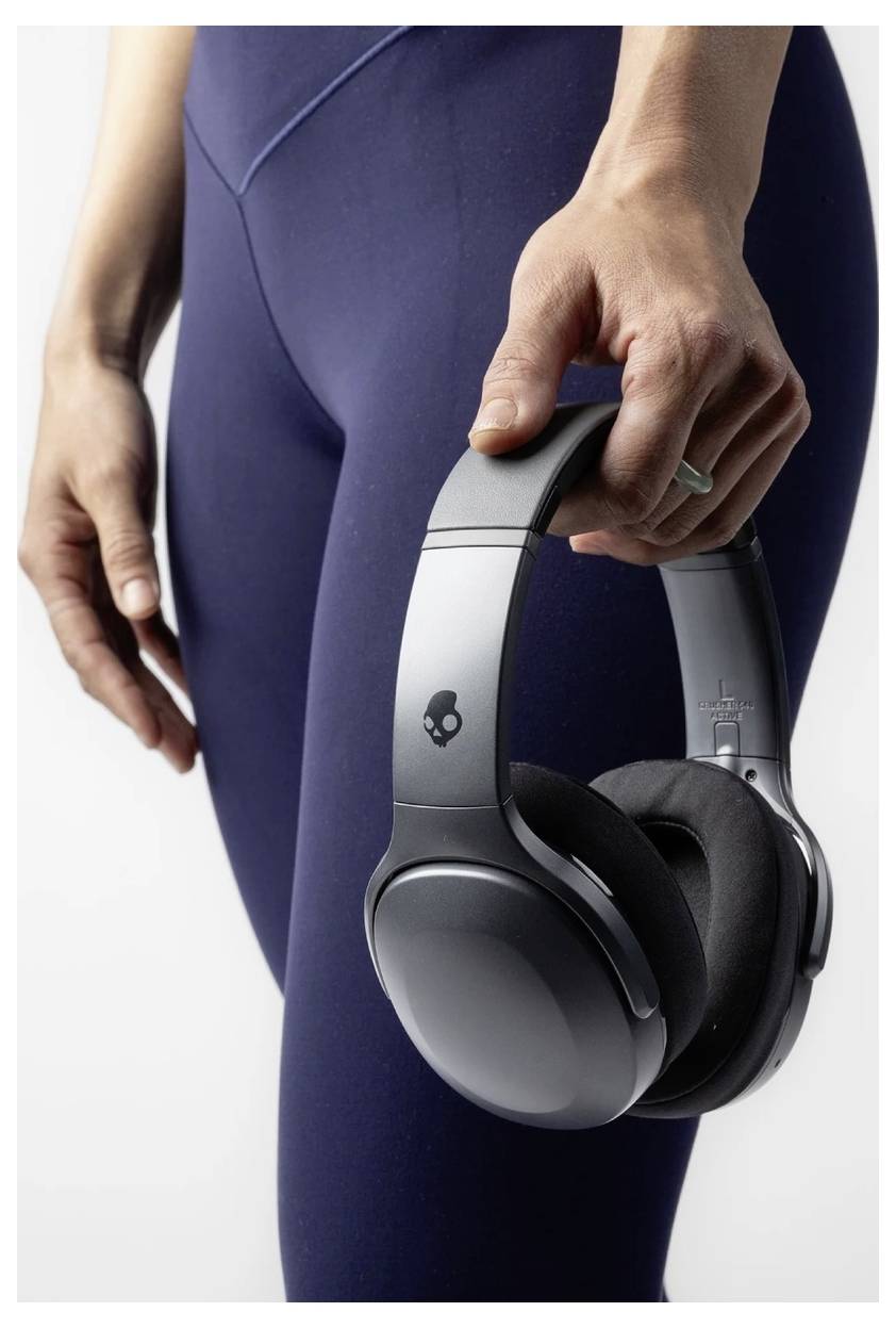 Une personne vêtue d'un pantalon bleu foncé tient des écouteurs gris supra-auriculaires dans sa main sur un fond clair.
