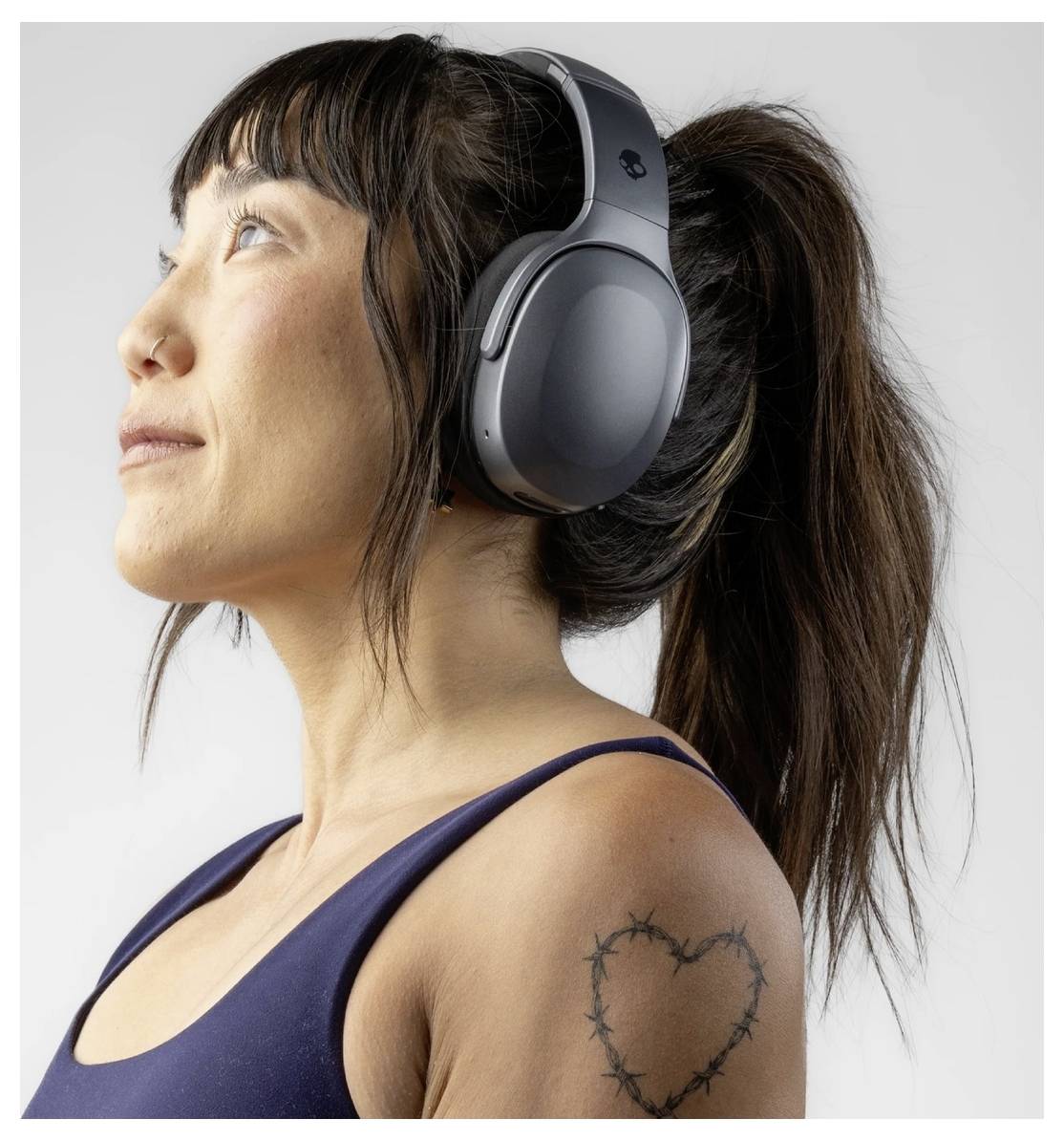 Une personne portant des écouteurs lève les yeux, vêtue d'un haut sans manches, et a un tatouage en forme de cœur entouré de fil barbelé sur l'épaule.