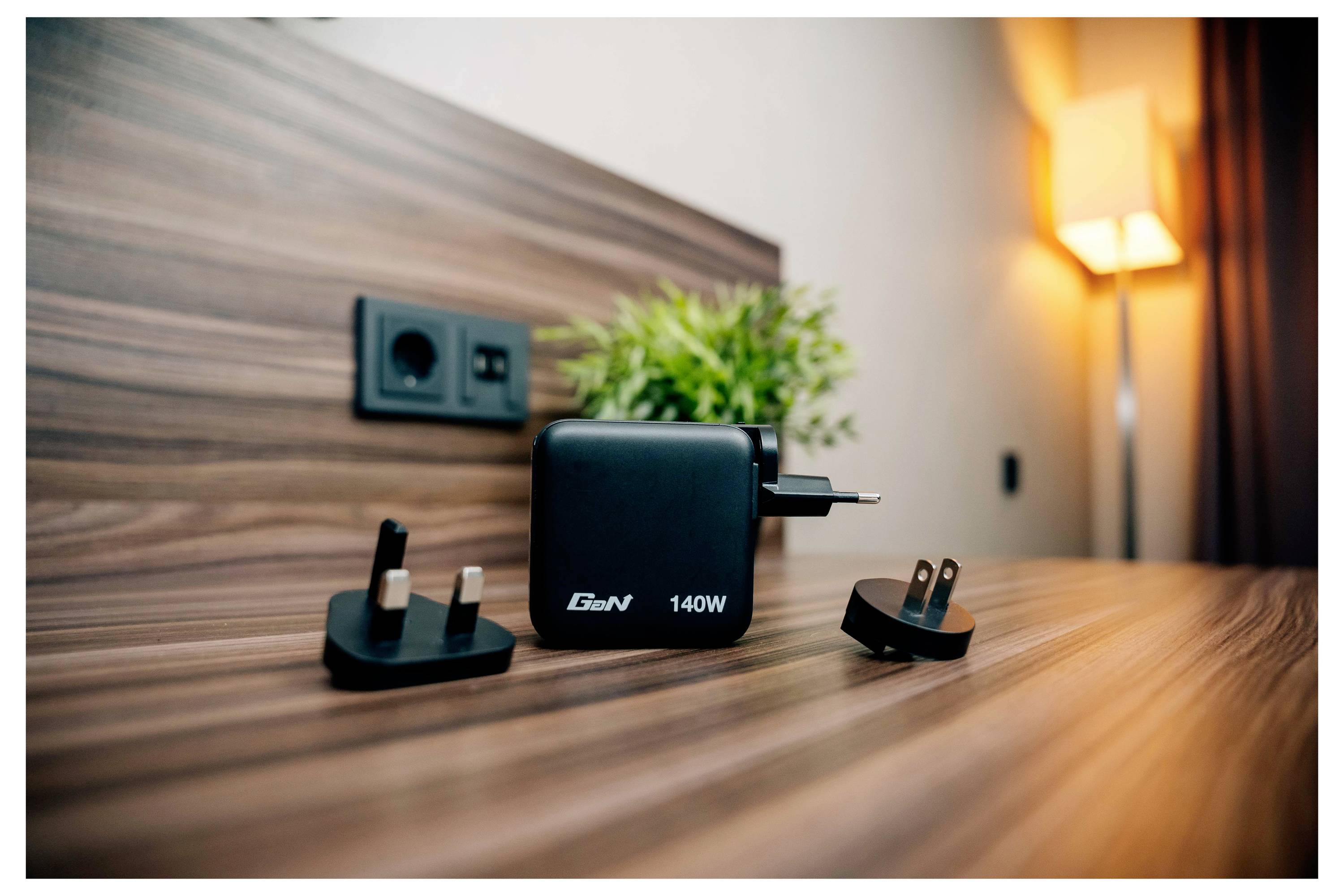 Un adaptateur de charge GaN noir 140W avec deux embouts de prise internationale repose sur une table en bois à côté d'une plante verte et d'une lampe.