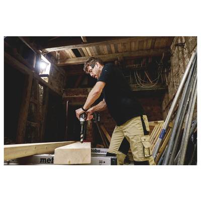 Un homme utilisant une perceuse électrique pour percer dans un bloc de bois dans un atelier ou un hangar de construction, avec des outils et du bois d'œuvre autour de lui.