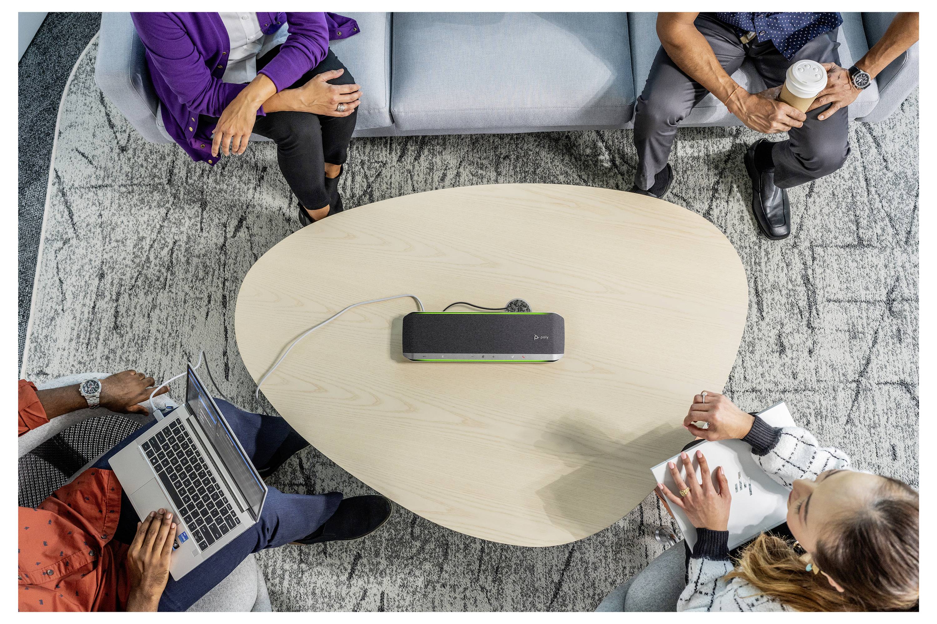 Quatre personnes assises autour d'une table ovale, collaborant avec des ordinateurs portables et des blocs-notes. Un appareil haut-parleur est placé au centre de la table.