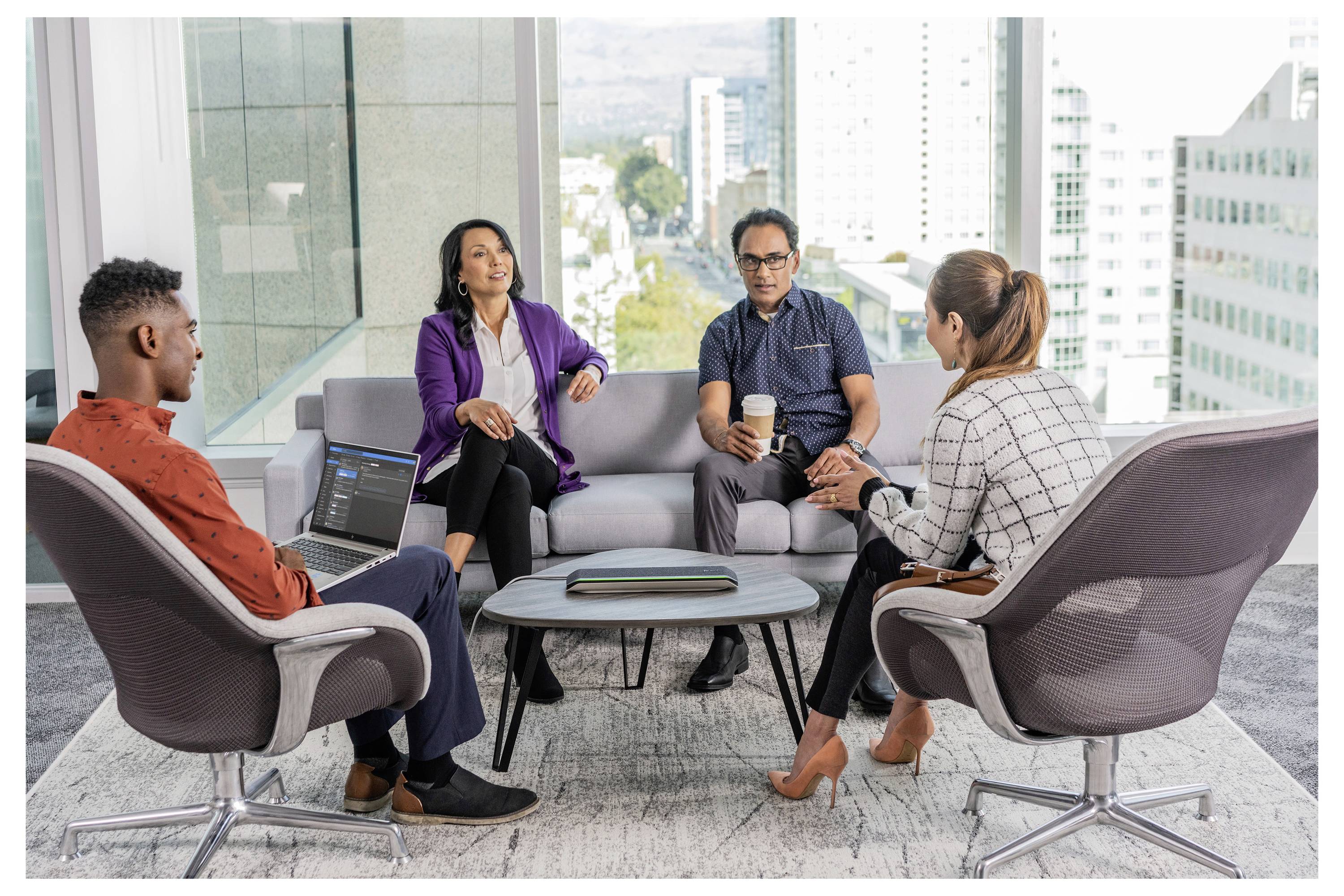 Quatre personnes engagées dans une réunion dans un cadre de bureau moderne, assises en cercle de chaises, avec un paysage urbain visible à travers de grandes fenêtres.