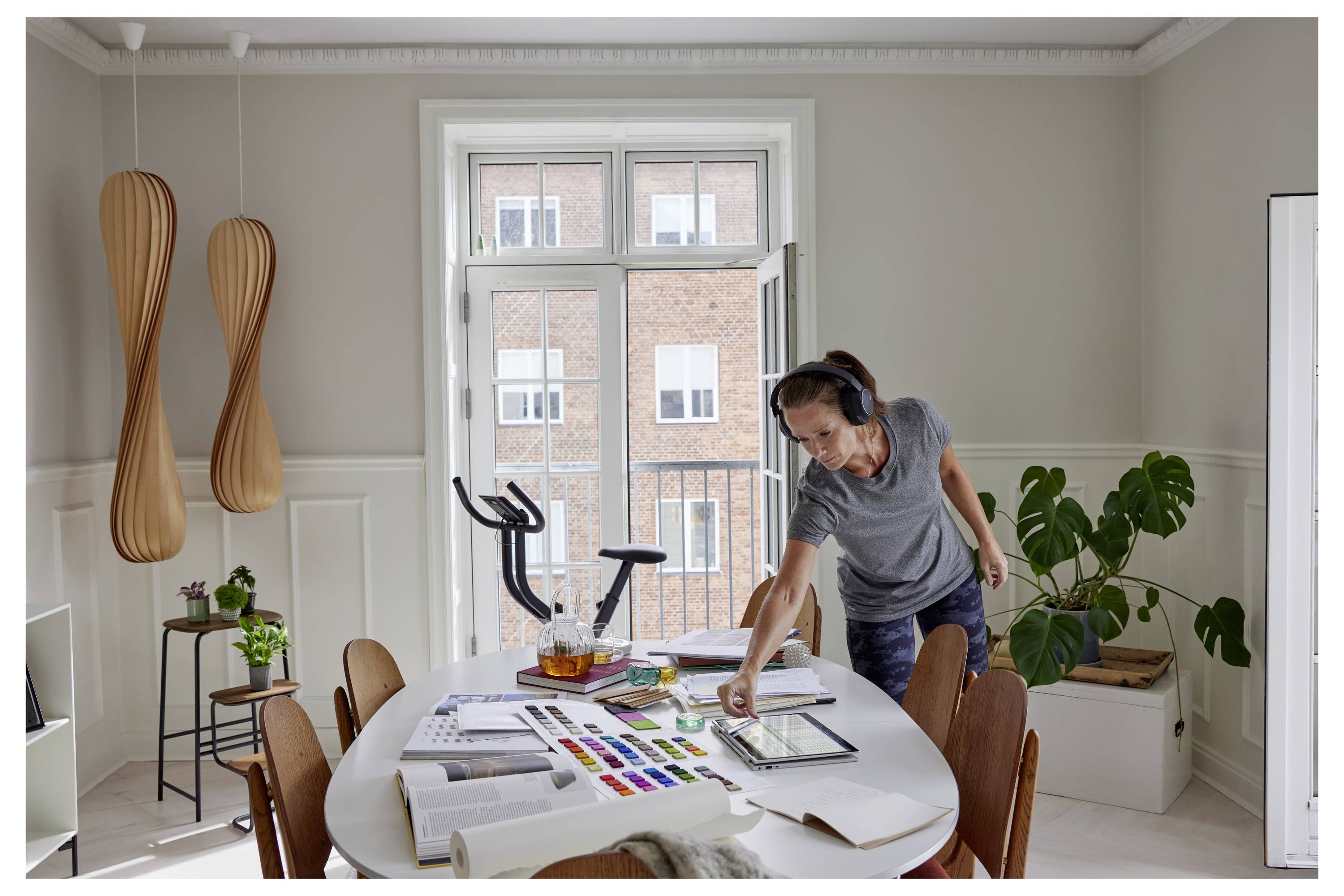 Une personne portant des écouteurs est dans une pièce lumineuse, sélectionnant des peintures sur une palette posée sur une table. Des équipements d'exercice et une plante sont visibles.