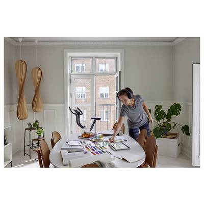 Une personne portant des écouteurs est dans une pièce lumineuse, sélectionnant des peintures sur une palette posée sur une table. Des équipements d'exercice et une plante sont visibles.