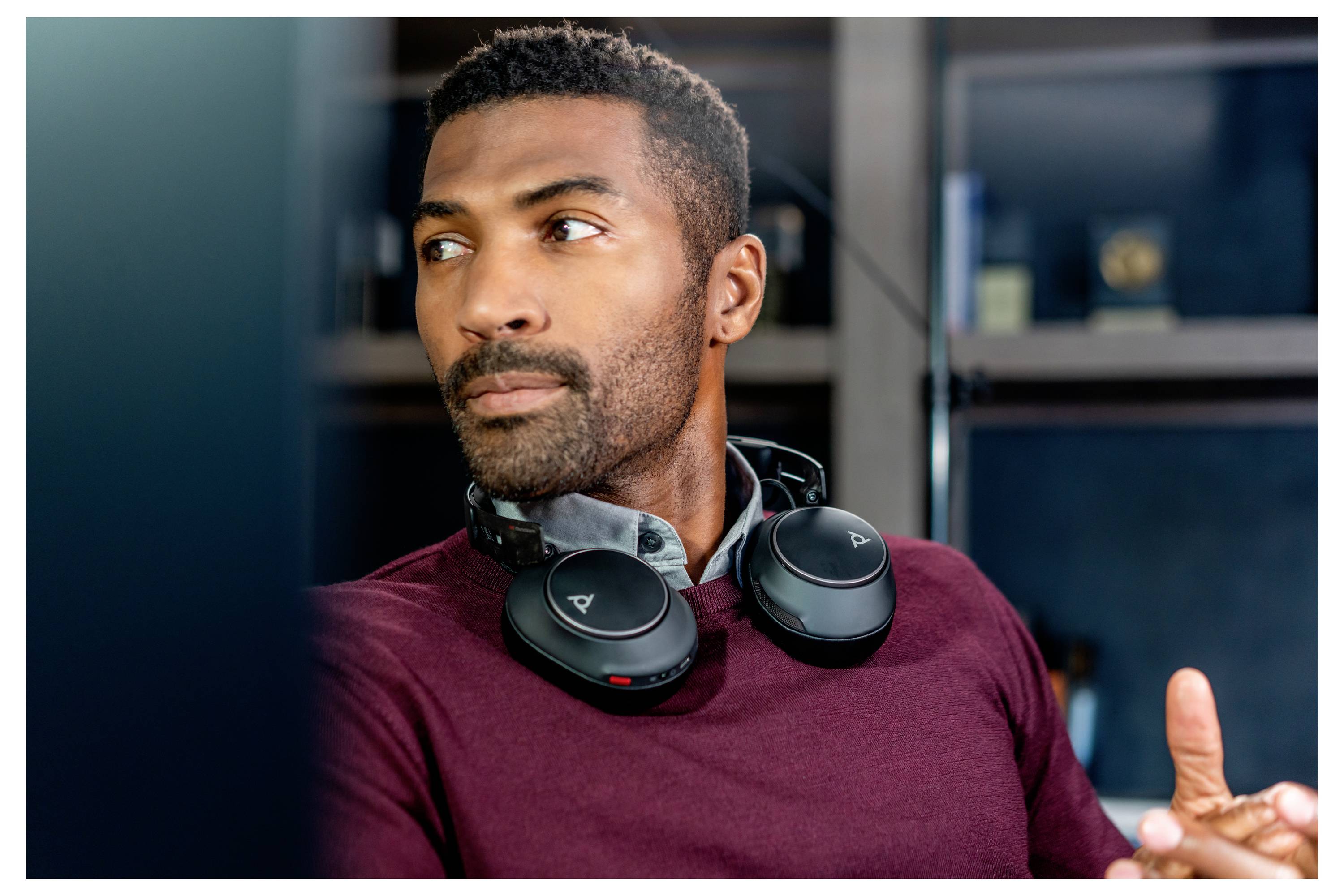 Un homme portant des écouteurs autour du cou est assis dans un bureau, regardant pensivement vers le côté. Des étagères remplies de livres sont visibles derrière lui.