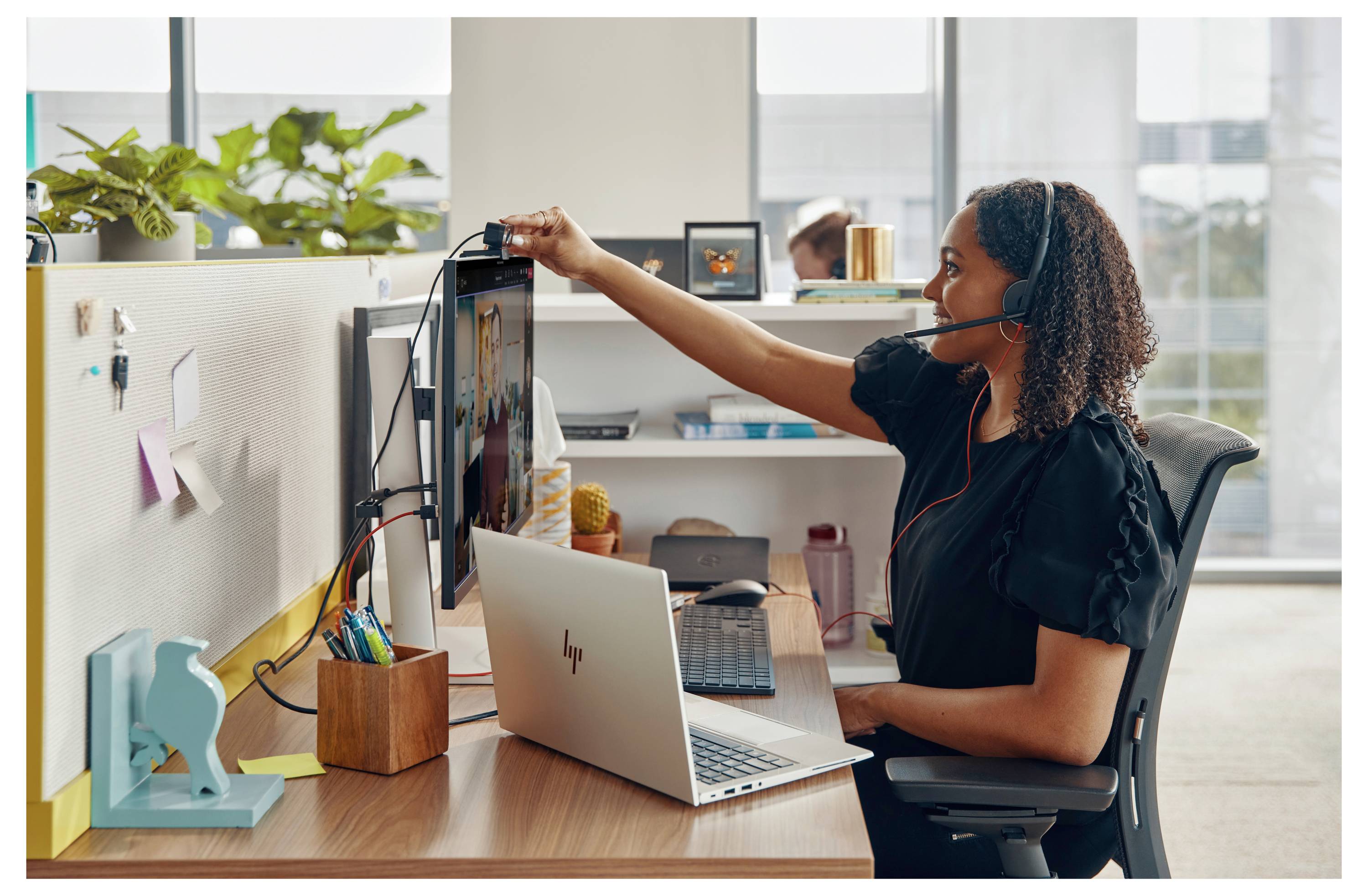 Une personne portant un casque travaille sur un ordinateur portable à un bureau dans un bureau, ajustant un moniteur vertical. L'espace de travail est organisé et décoré.
