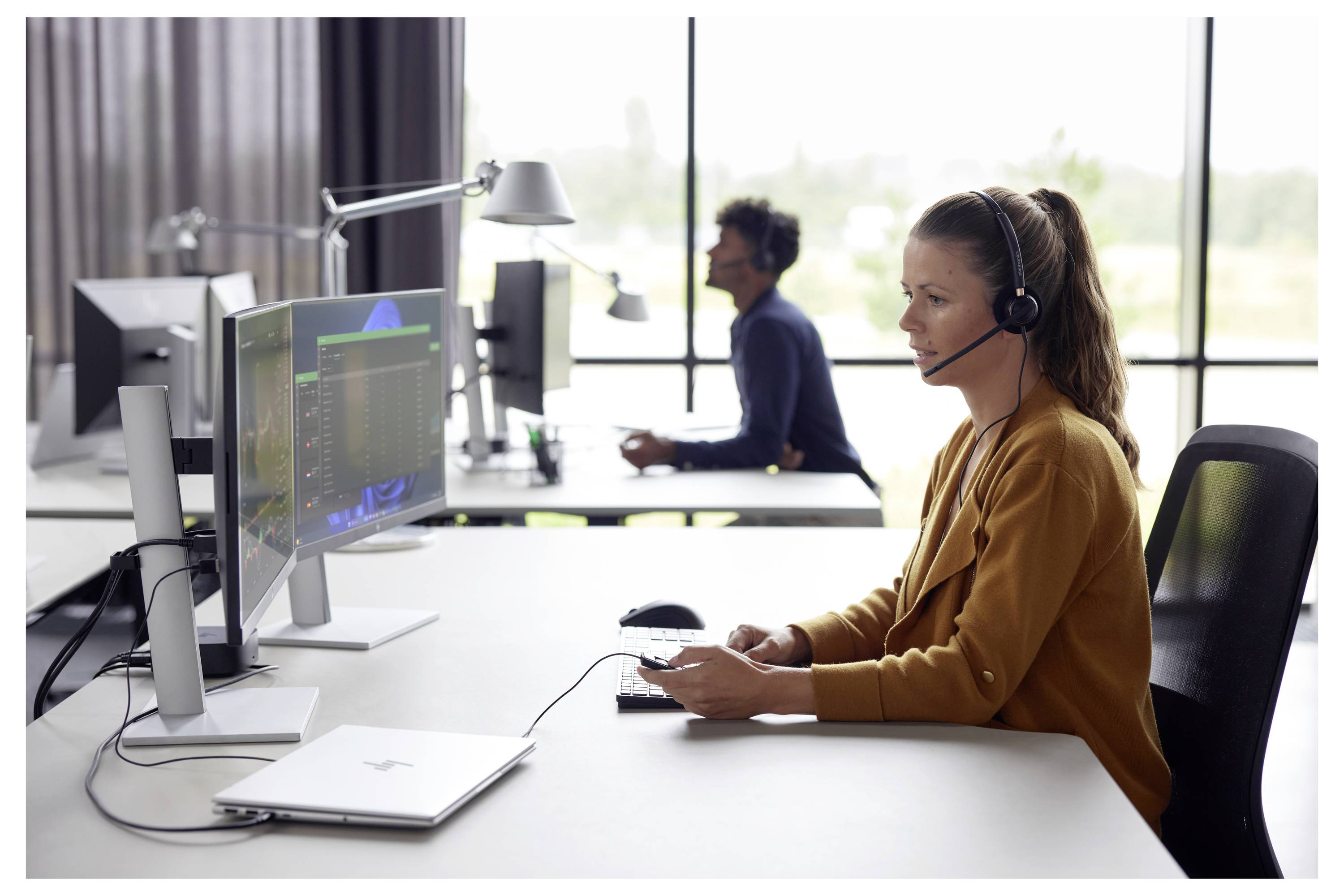 Femme portant un casque à un poste de travail informatique dans un centre d'appels, avec un autre utilisateur de casque travaillant en arrière-plan.