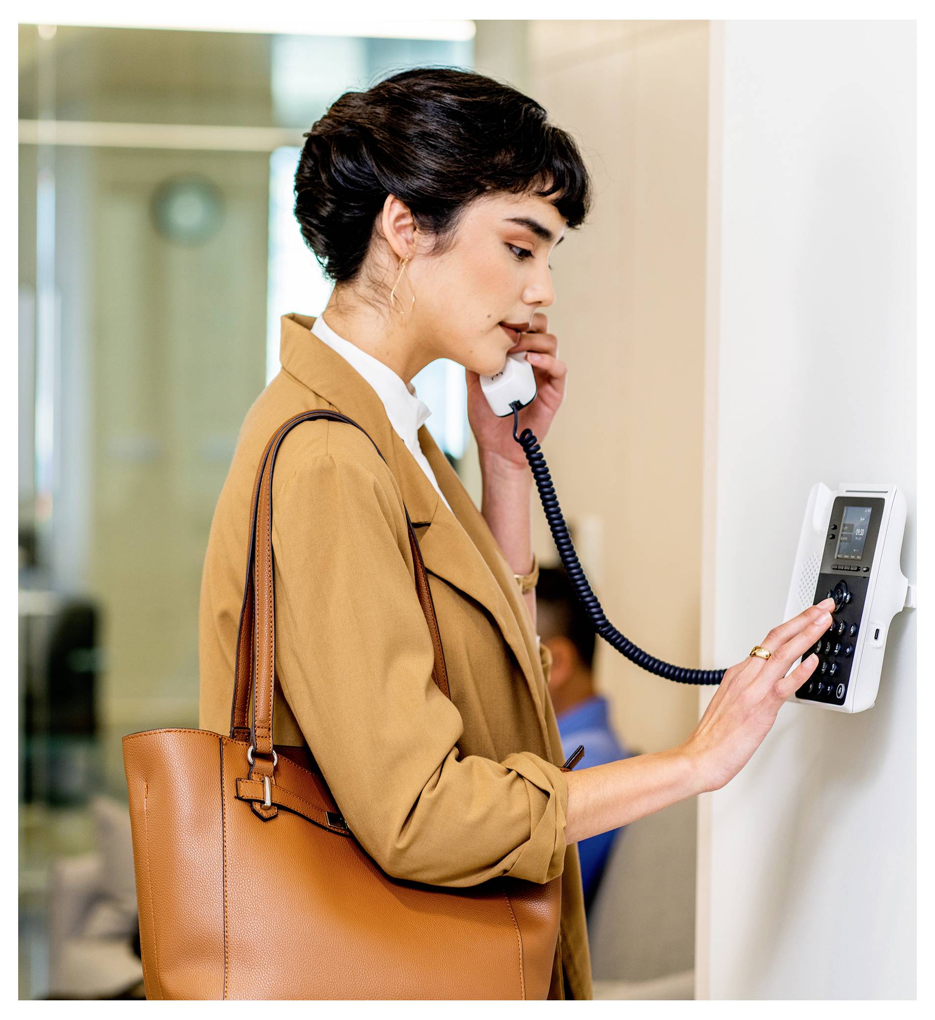 Une femme dans un manteau beige utilise un interphone mural en parlant dans le combiné, avec un sac à bandoulière marron sur son bras.