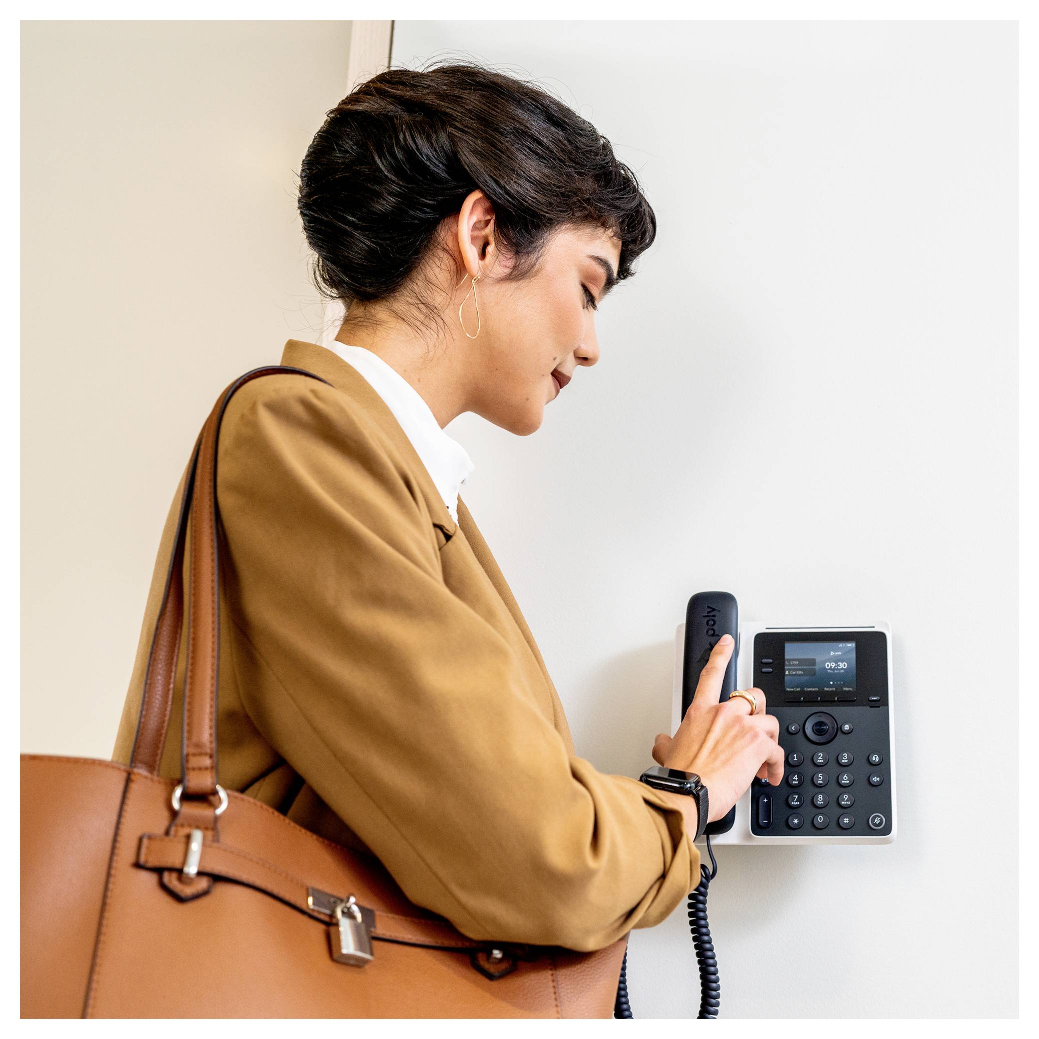 Femme vêtue d'un manteau beige et d'un sac à main utilise un clavier de téléphone de bureau monté au mur, appuyant sur un bouton tout en regardant l'écran.