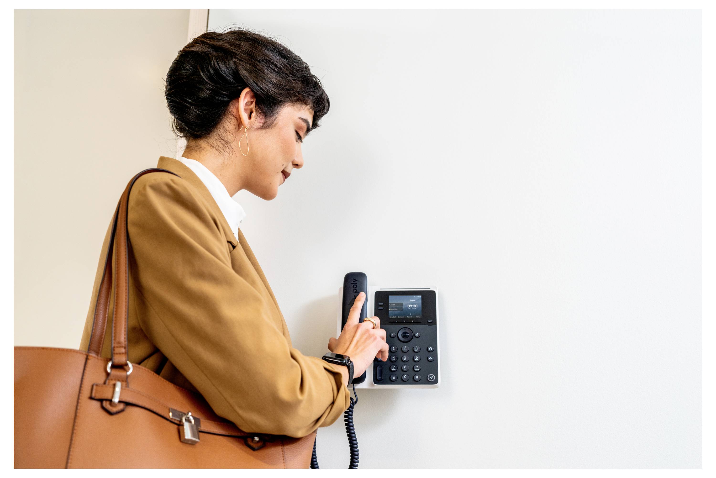 Une femme dans un manteau beige utilise un téléphone de bureau mural ou un interphone, tenant le combiné à côté d'un mur blanc vierge.