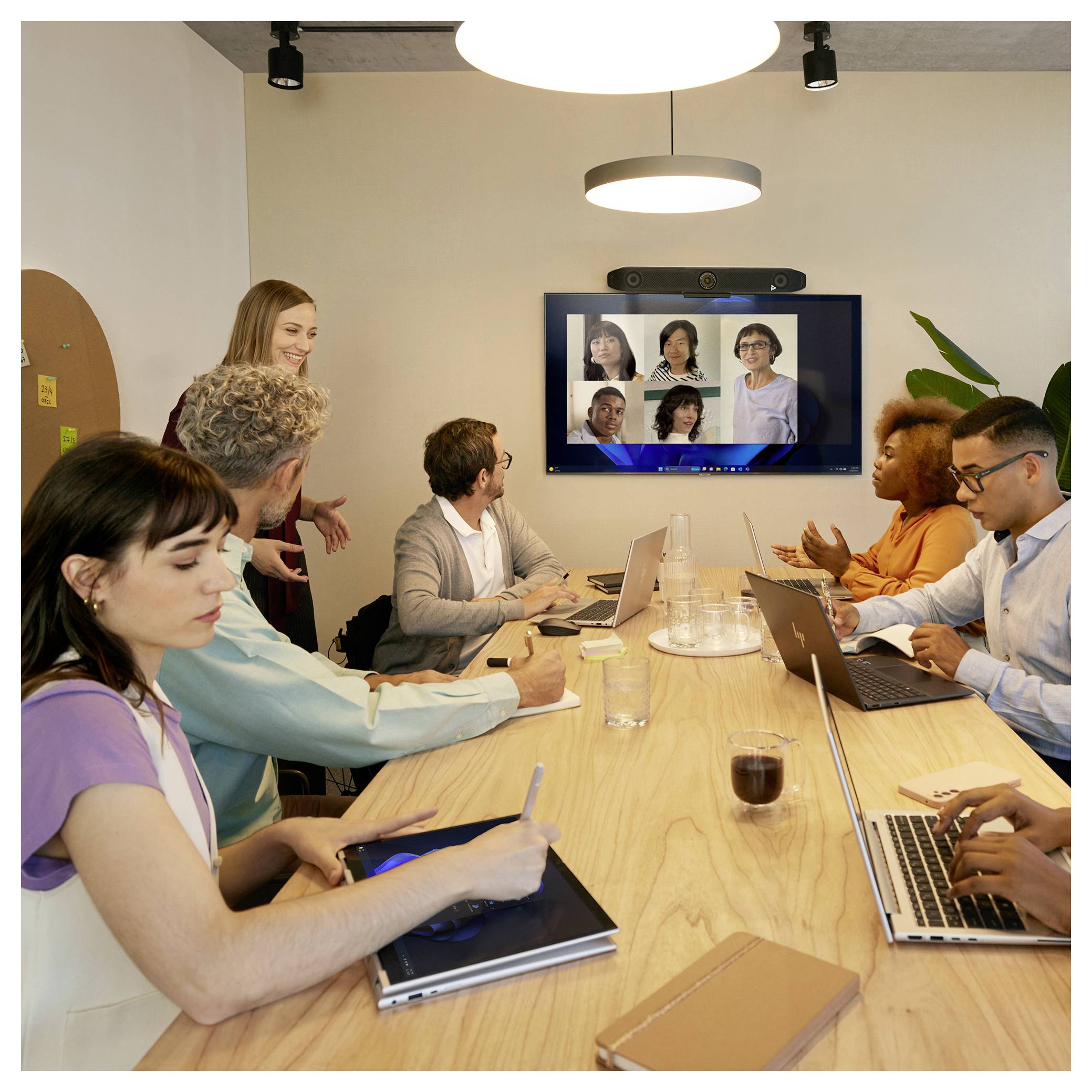 Réunion d'équipe autour d'une table de conférence avec plusieurs personnes utilisant des ordinateurs portables et regardant un écran mural affichant un appel vidéo à distance avec cinq participants.