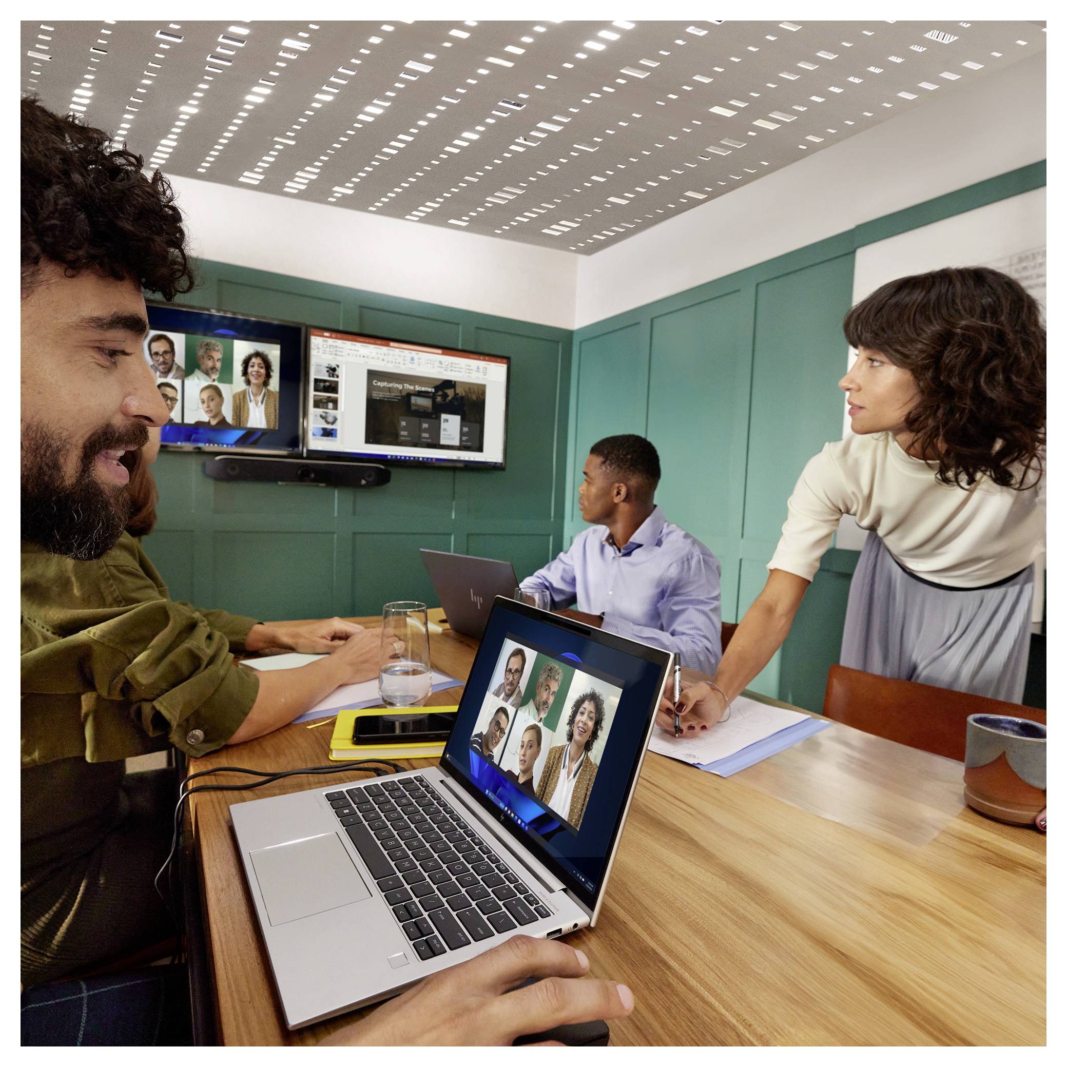 Réunion d'équipe dans une salle de conférence avec un appel vidéo sur des ordinateurs portables et des écrans muraux ; quatre collègues collaborent autour d'une table.