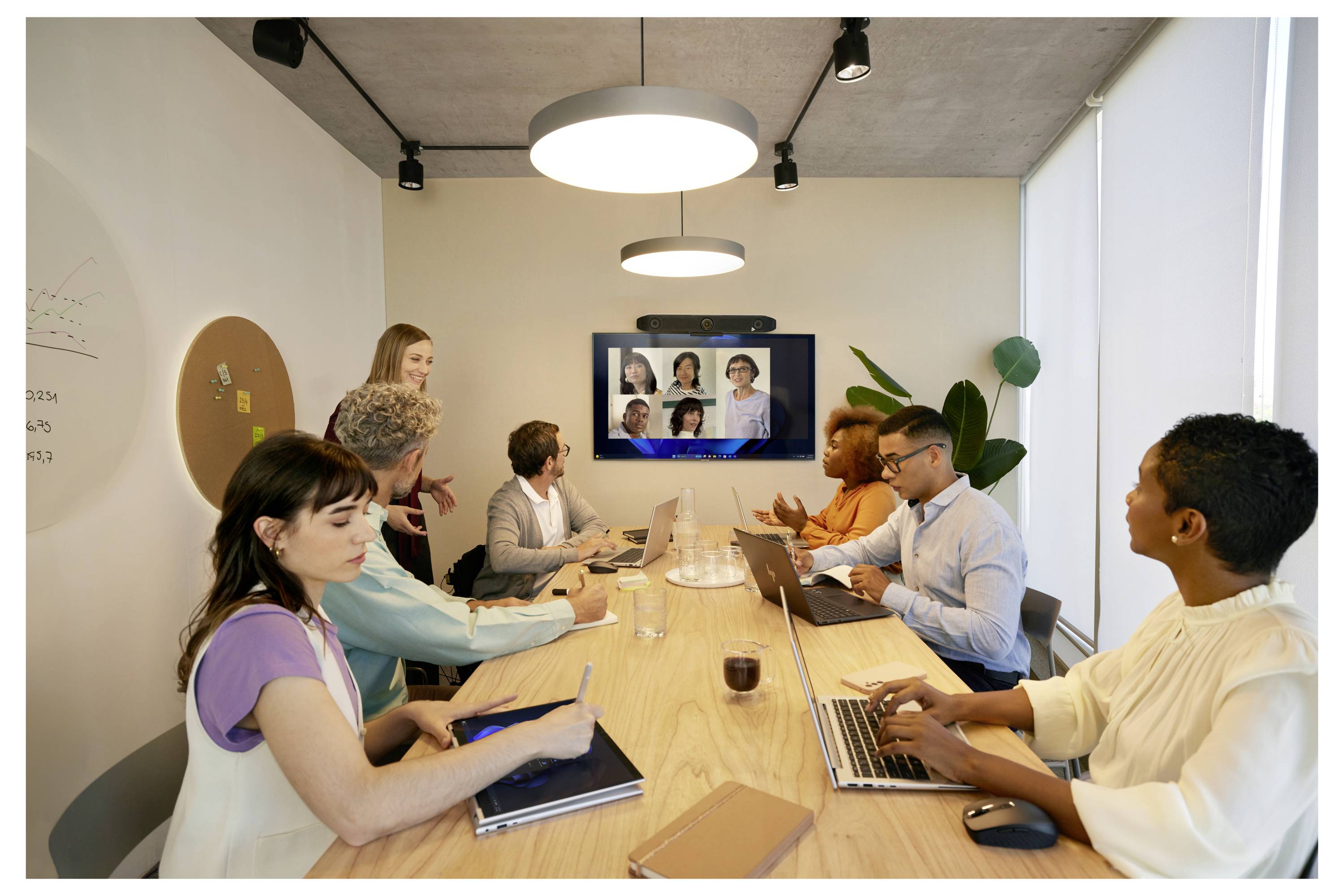 Des personnes dans une salle de conférence se réunissent autour d'une table en utilisant des ordinateurs portables pour un appel vidéo affiché sur un écran mural.