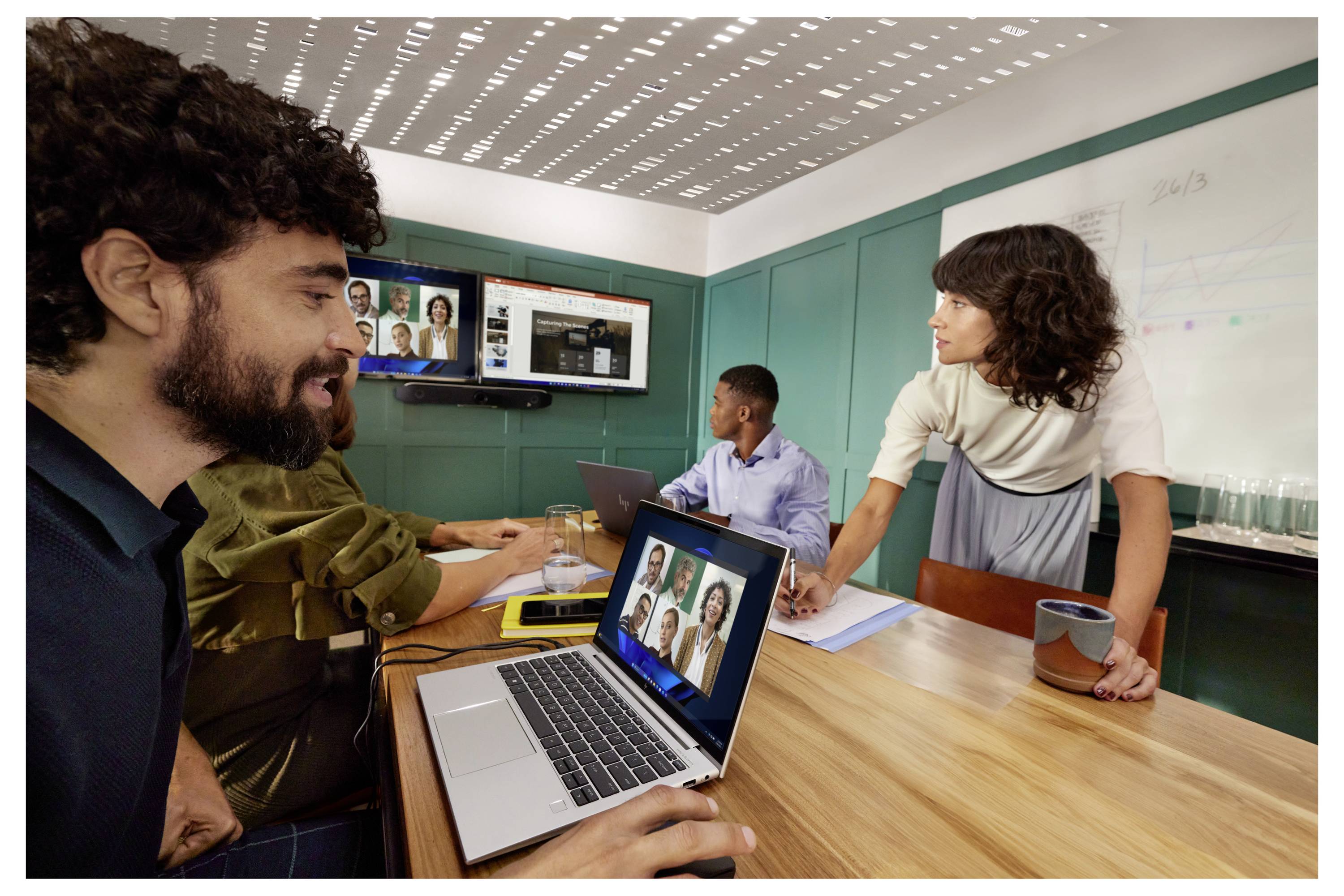 Des personnes dans une salle de réunion collaborent autour d'une table de conférence avec des ordinateurs portables et un écran mural affichant un appel vidéo.