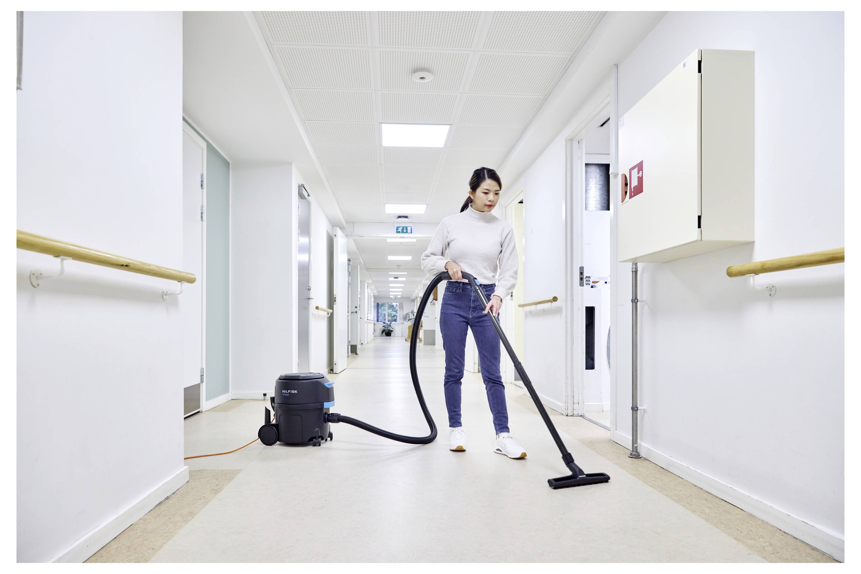 Une personne passant l'aspirateur dans un couloir aux murs blancs et aux portes blanches, utilisant un aspirateur noir. Le sol est beige, et il y a des rampes d'appui sur les murs.