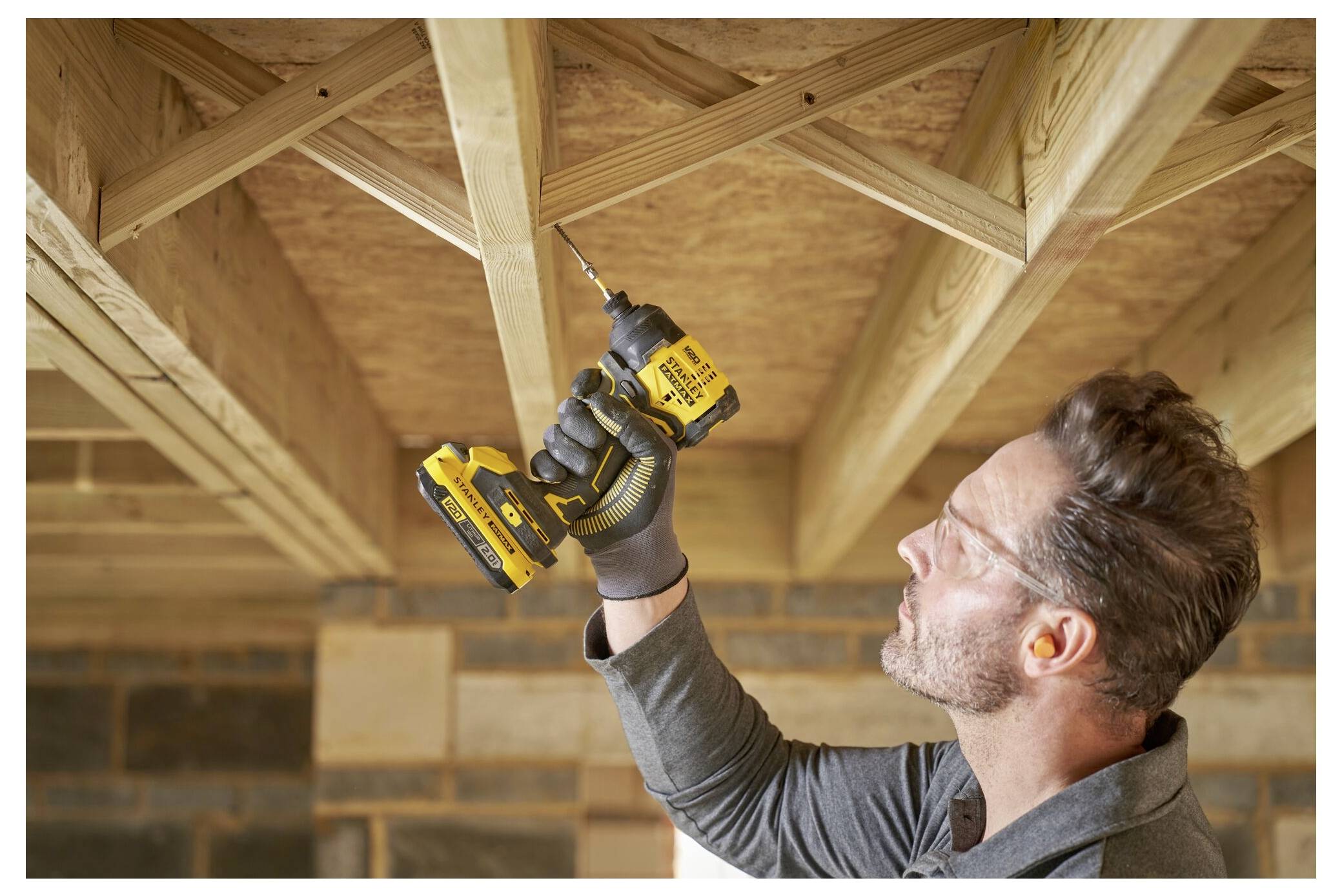 Une personne installe une charpente de plafond en bois à l'aide d'une perceuse-visseuse jaune et noire dans une pièce en construction.