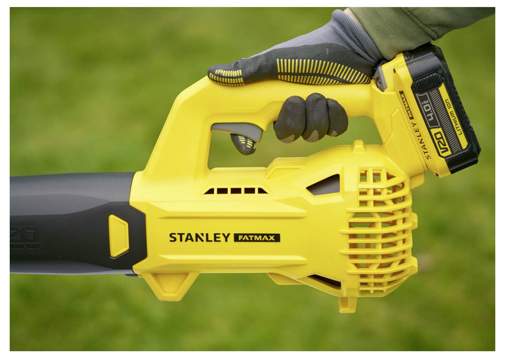 Souffleur de feuilles « Stanley FatMax » tenu par une main gantée, mettant en évidence la conception et le compartiment de batterie sur un fond extérieur vert.