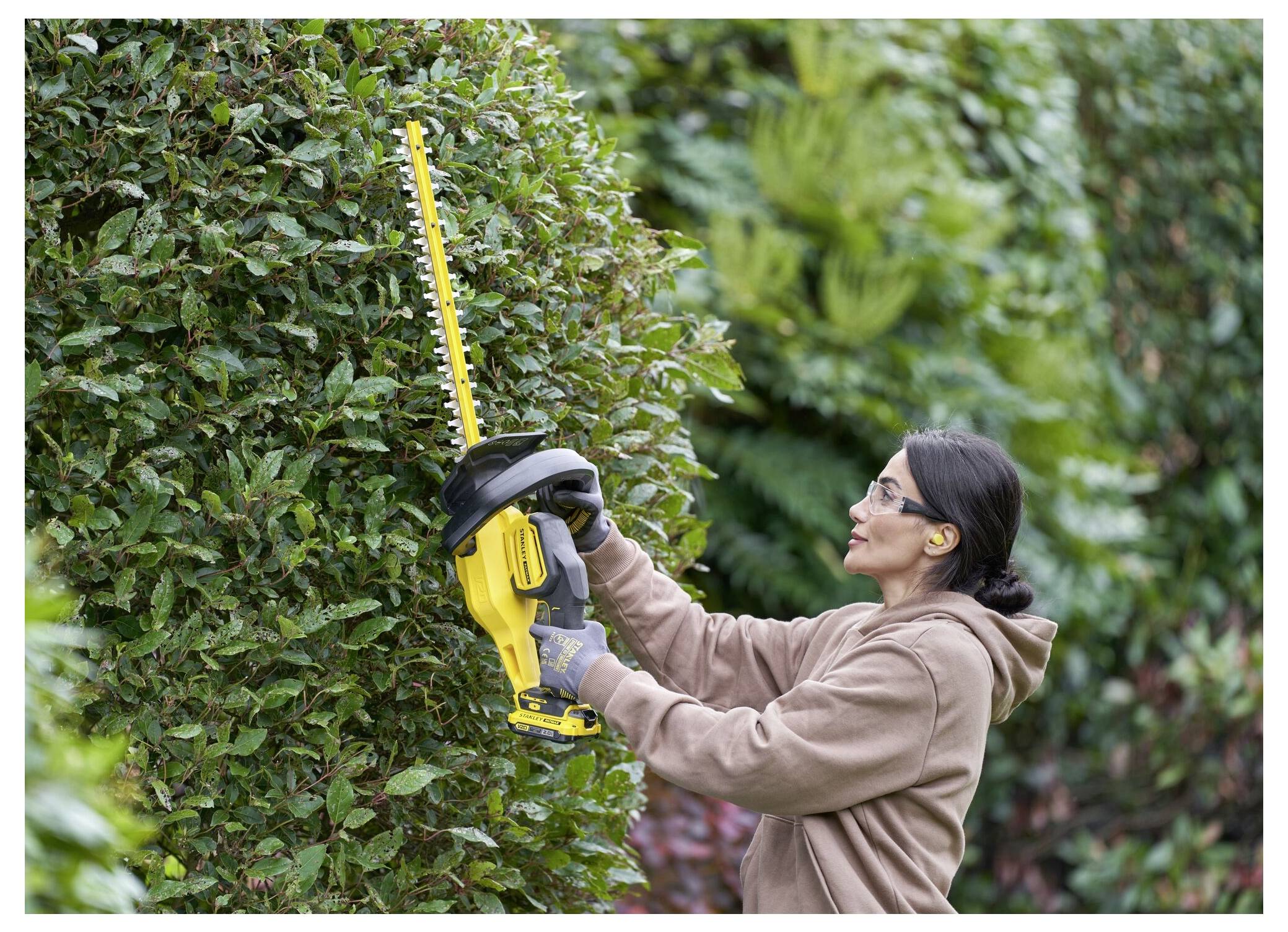 Une personne portant des lunettes de protection et des gants utilise un taille-haie électrique pour façonner un grand buisson dans un jardin.