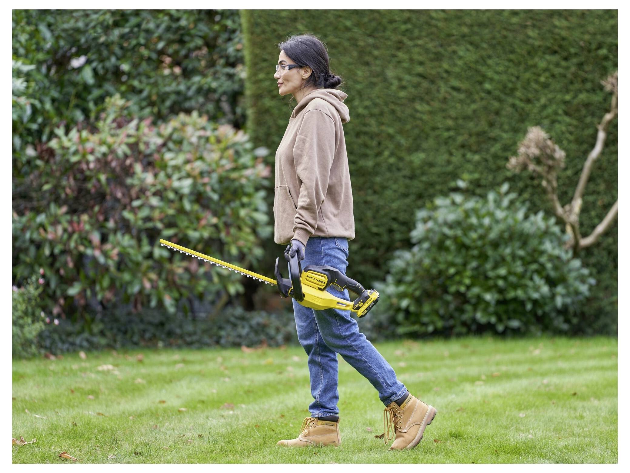 Une personne taillant une haie dans un jardin avec un taille-haie électrique jaune, portant un sweat-shirt à capuche marron, un jean, des gants et des bottes.