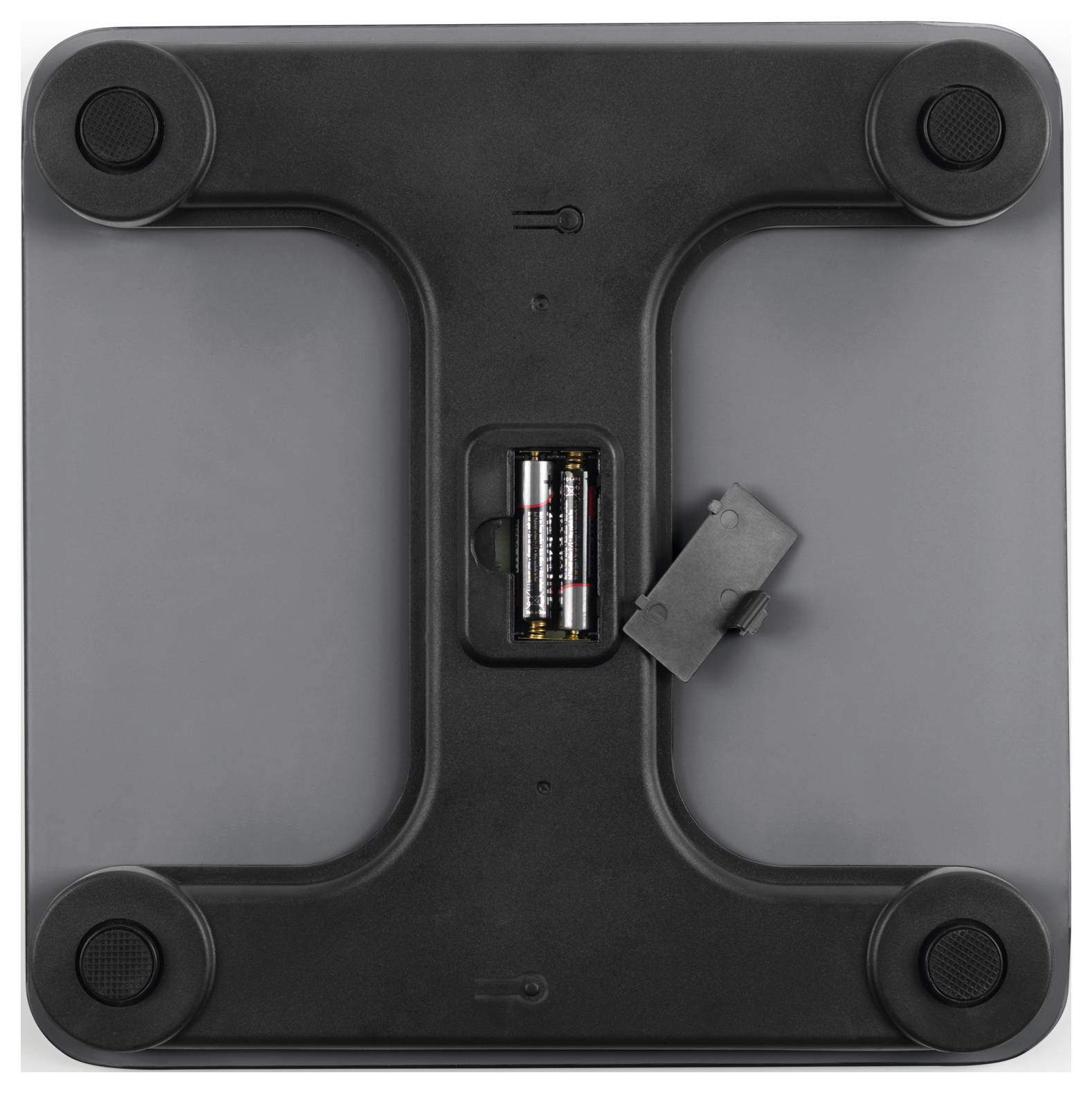 L'image montre le dessous d'un appareil électronique, probablement une balance, avec des batteries installées dans un compartiment et quatre pieds en caoutchouc.