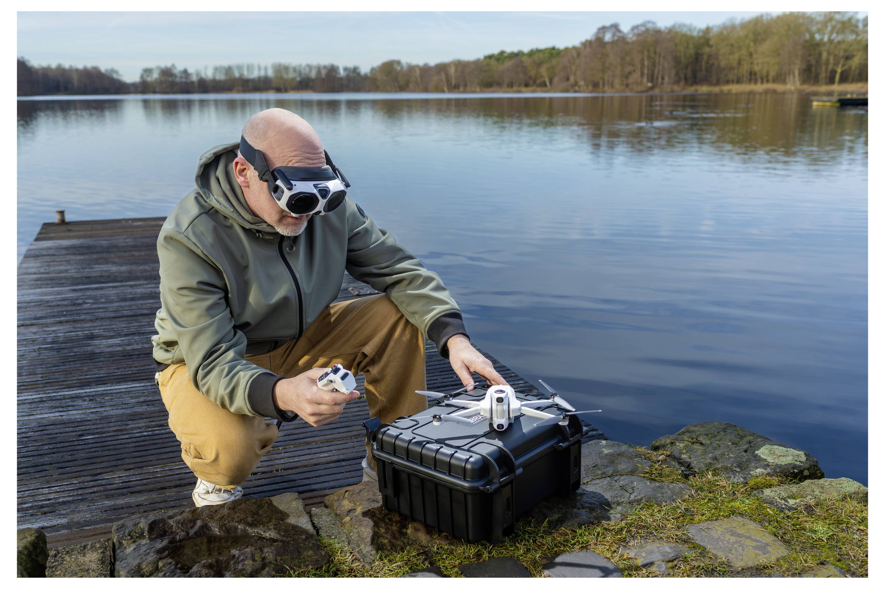 Une personne accroupie près d'un quai au bord d'un lac pilote un drone avec une télécommande, portant des lunettes de vision à la première personne pour la navigation.