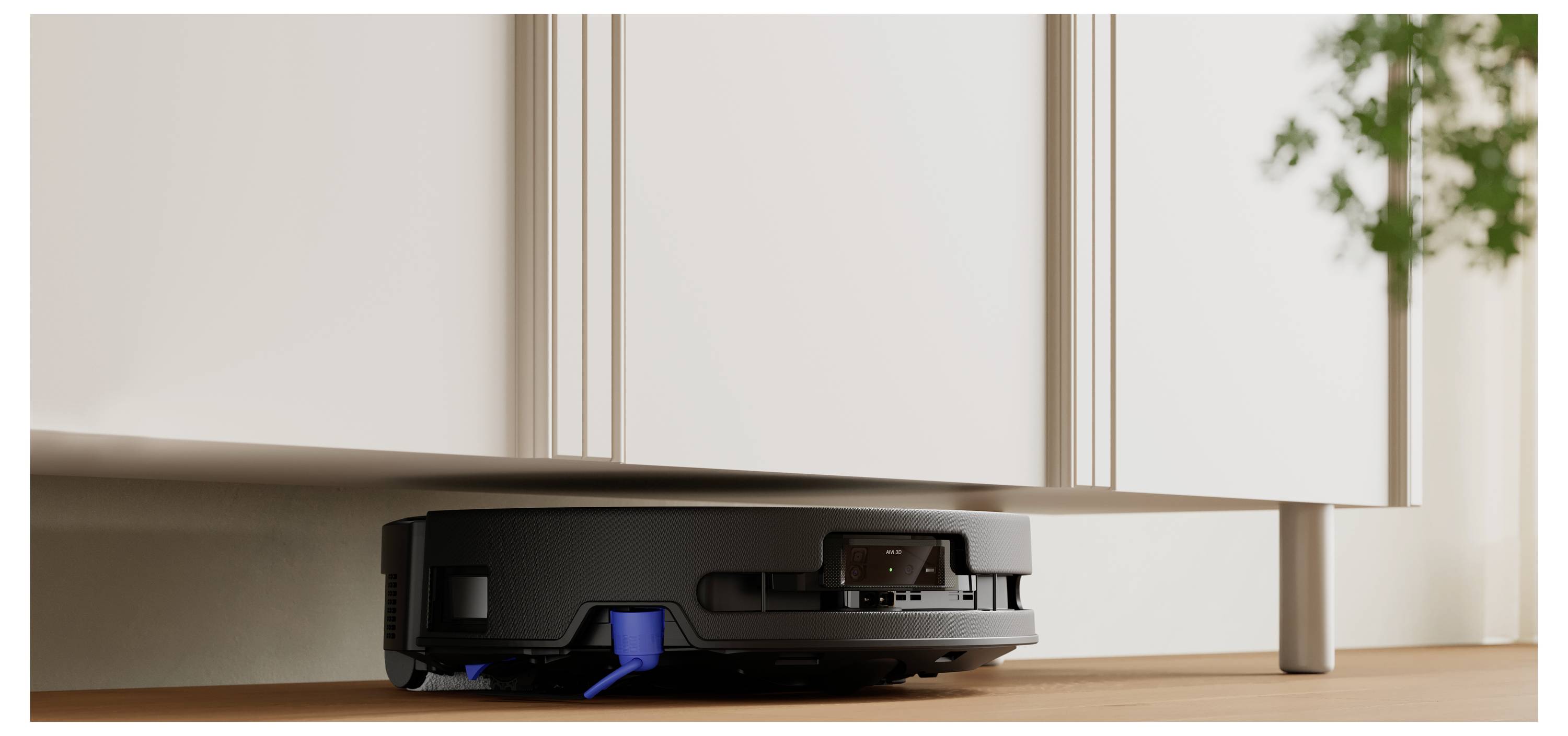 Un aspirateur robot noir navigue sous un meuble blanc sur un sol en bois, avec une plante visible en arrière-plan.
