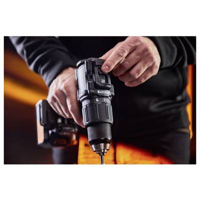 Une personne utilisant une perceuse sans fil, tenant et serrant un foret, avec un bloc de batterie attaché et des vêtements de travail orange en arrière-plan.