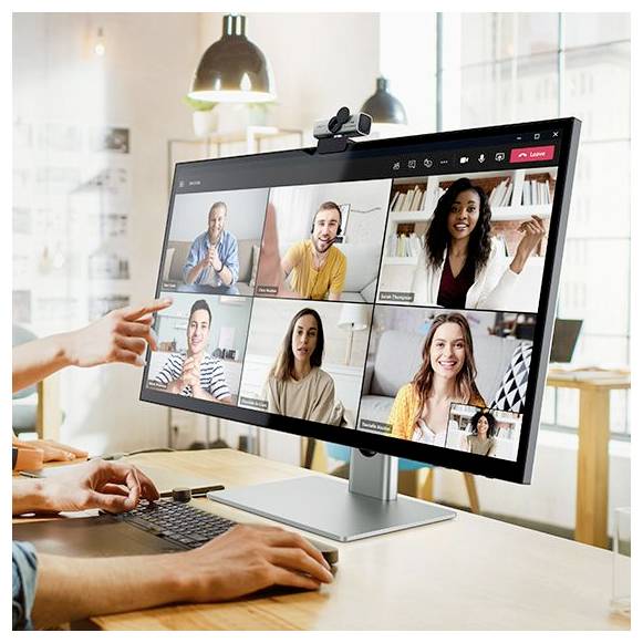 Une personne participe à une visioconférence sur un moniteur de bureau affichant six participants souriants dans une grille, avec une webcaméra attachée et un clavier sur le bureau.