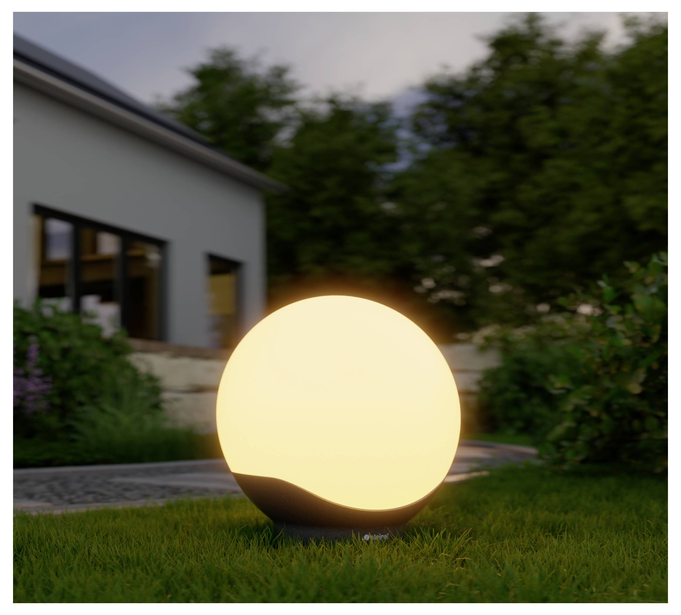 Lampe extérieure sphérique brillante sur l'herbe dans un jardin au crépuscule, à côté d'une maison moderne et d'un mur en pierre.