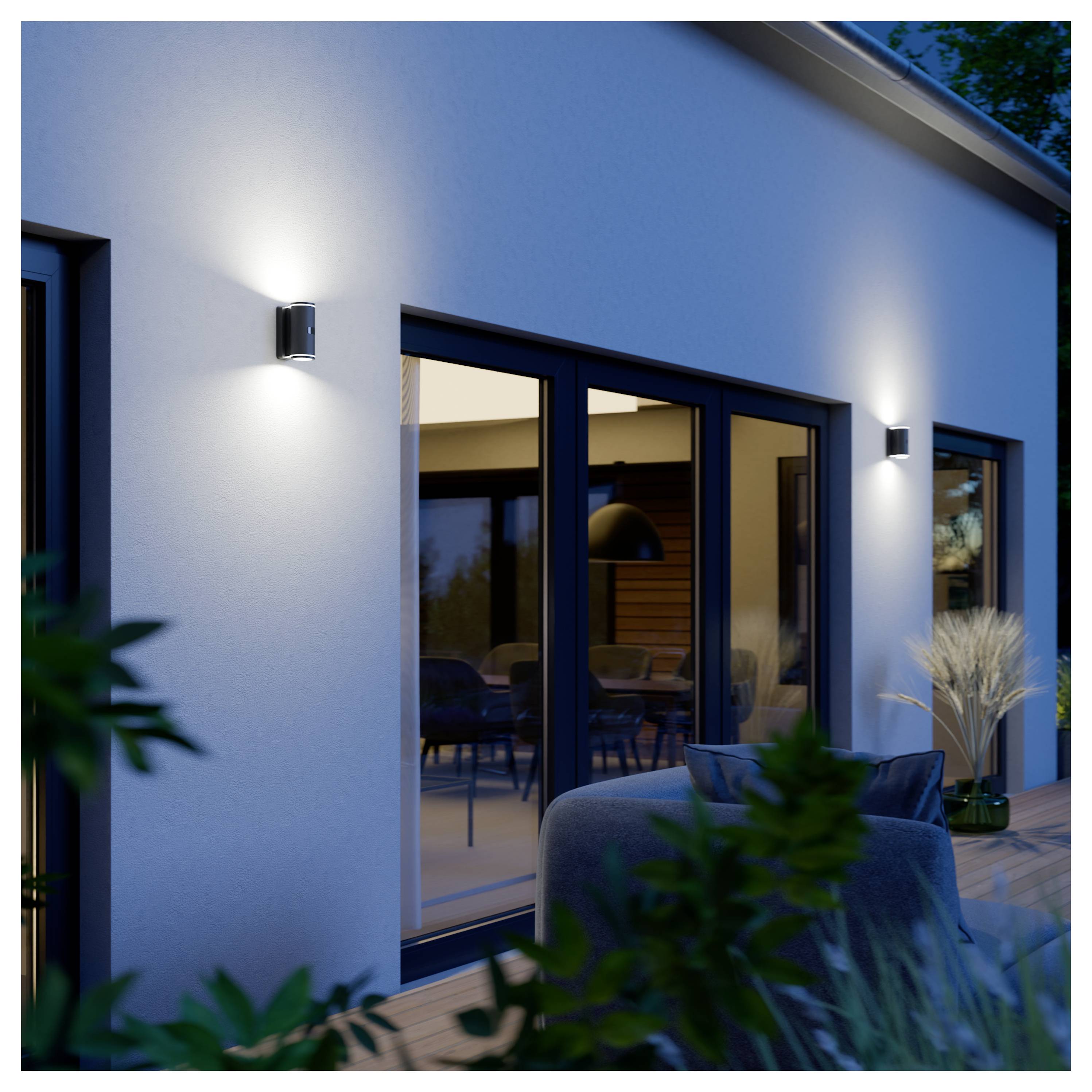 Extérieur de maison moderne au crépuscule avec appliques murales éclairées, grandes portes coulissantes en verre, et une terrasse avec mobilier et plantes.